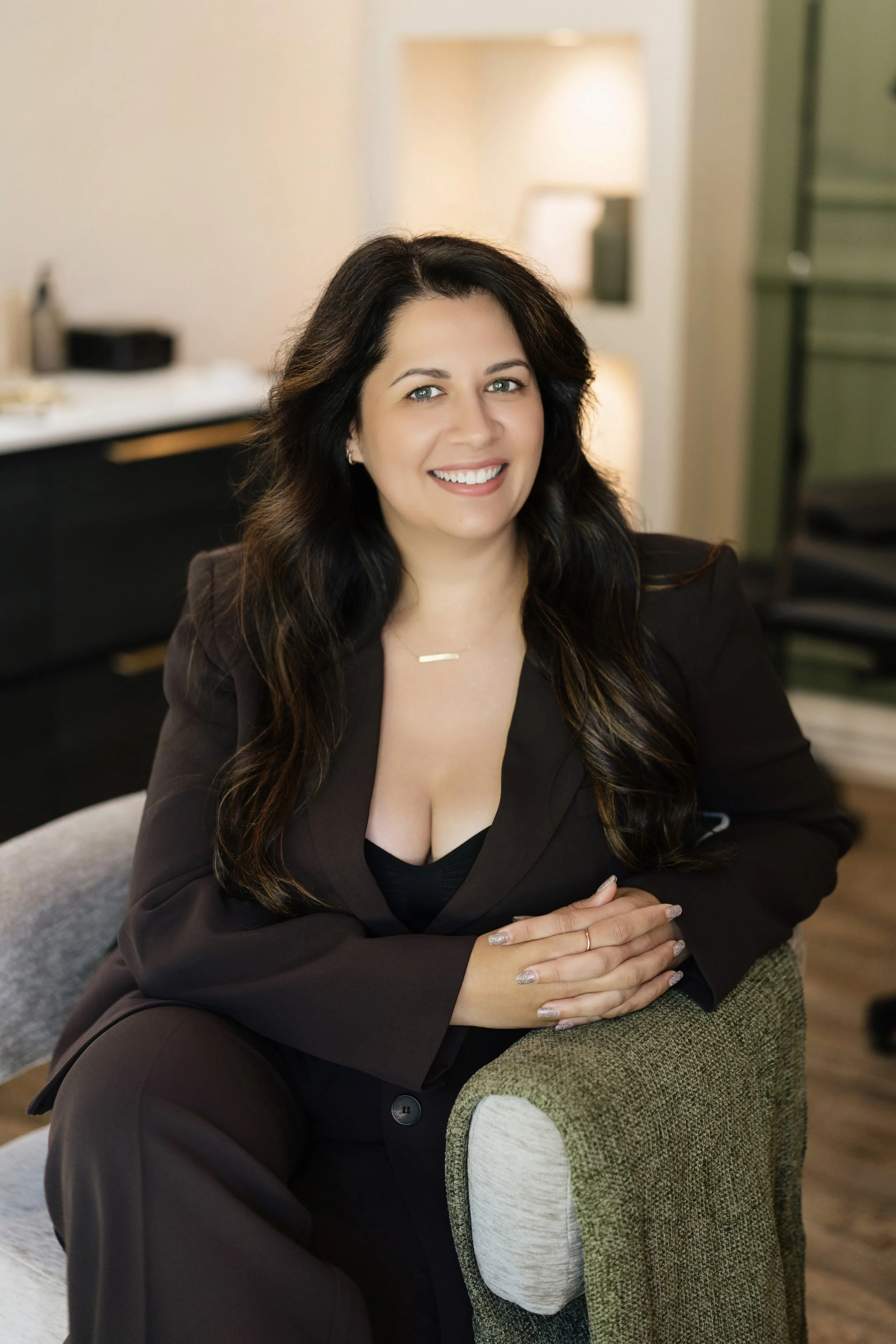 A woman with long dark hair smiling, sitting at a desk with hands folded, in an indoor setting with a lamp and furniture in the background.