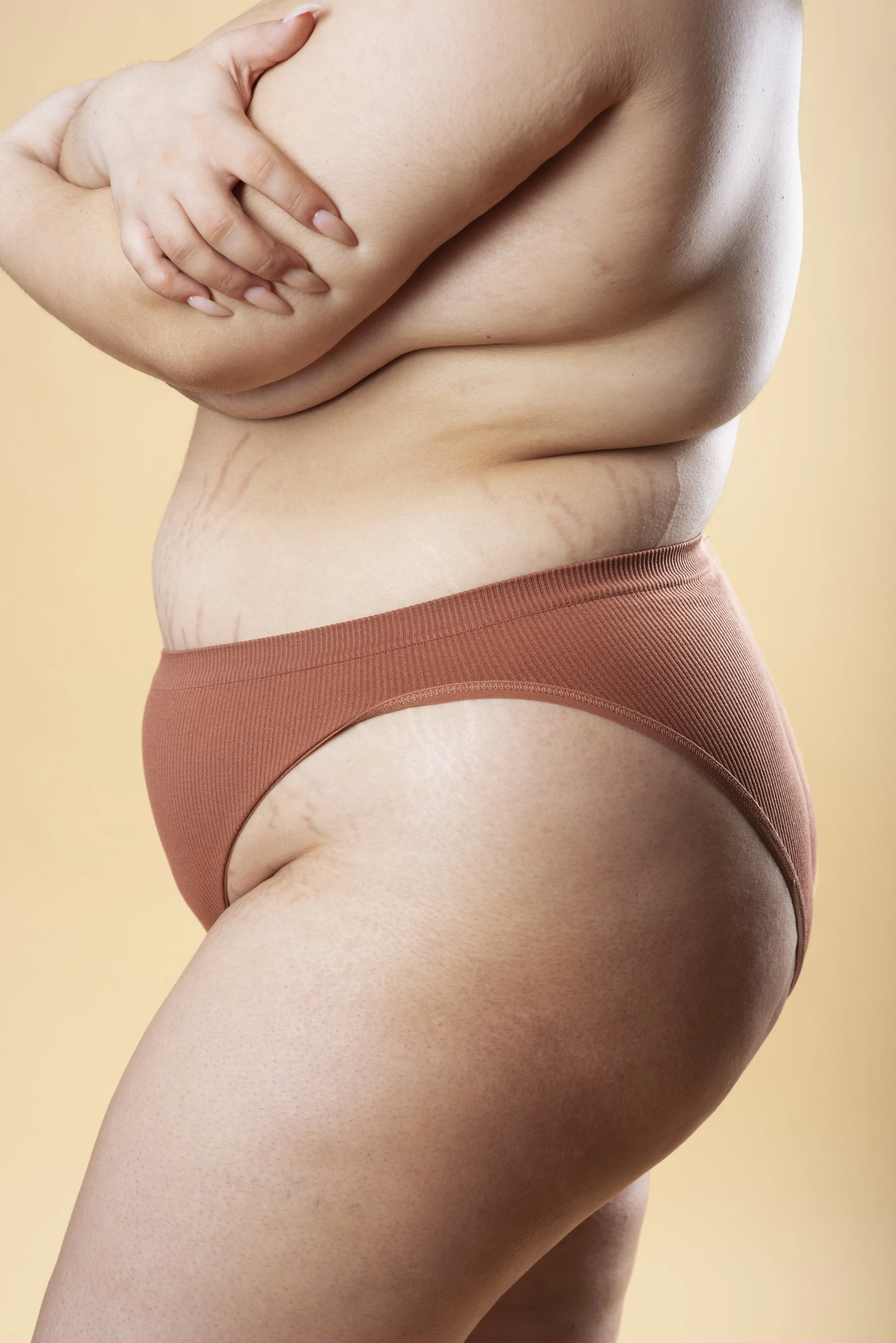 Close-up of a woman's midsection with visible stretch marks, wearing a brown thong, with her hand on her upper arm against a beige background.