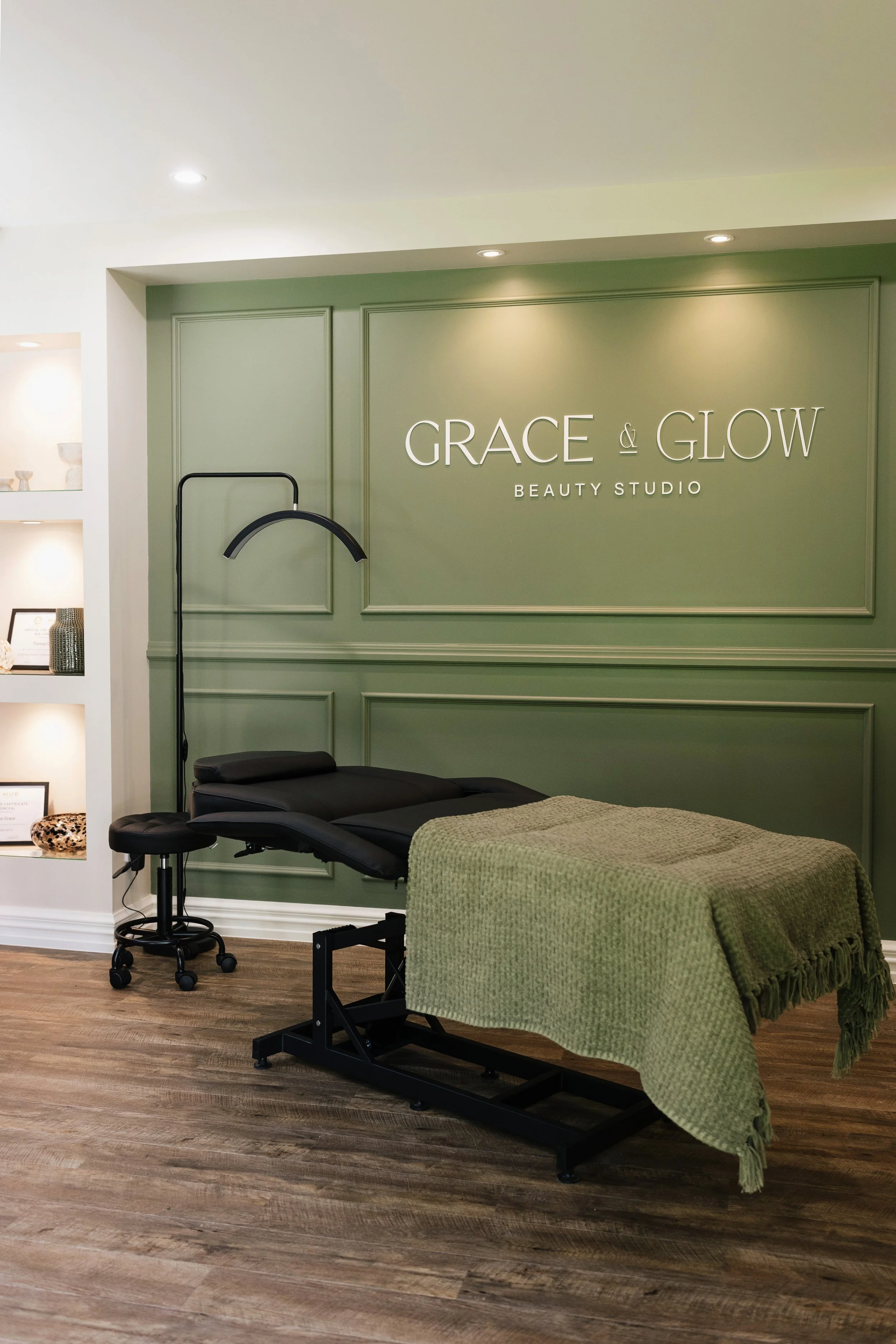 Interior of a beauty studio with a massage bed covered in a green blanket, a black lamp, and a wall sign that reads 'GRACE & GLOW BEAUTY STUDIO'.