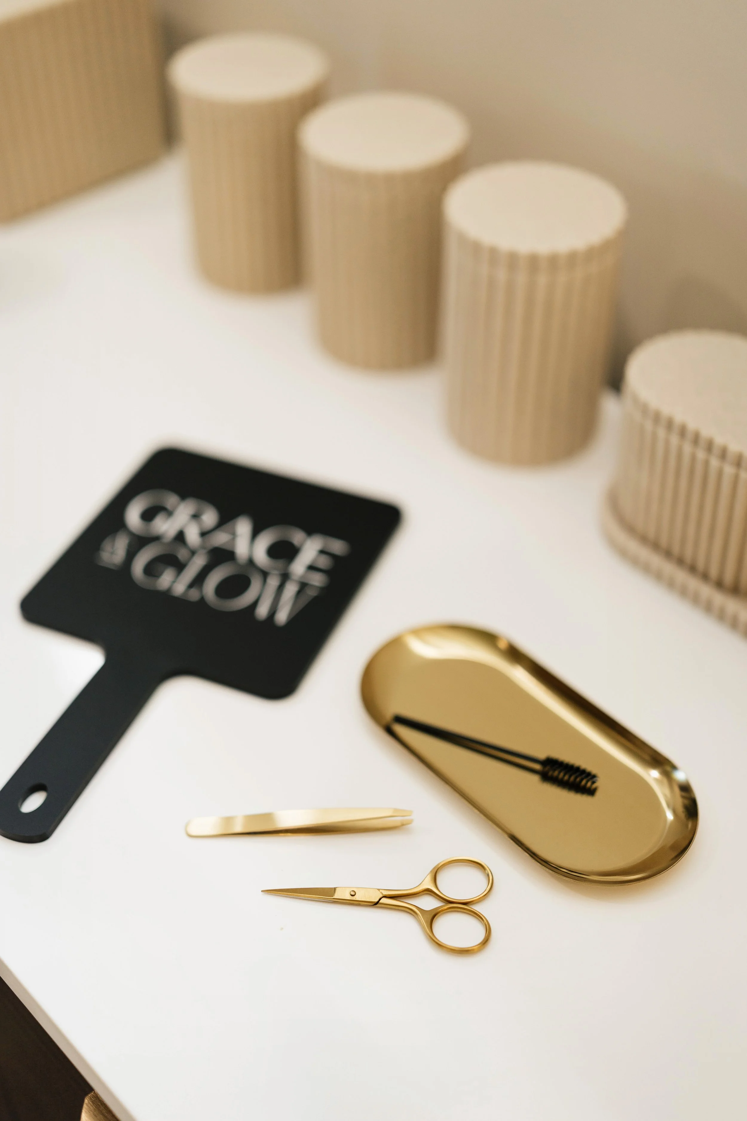 A table with a gold tray, gold scissors, a gold tweezer, and a sign that says 'Grace & Grit', with beige corrugated cardboard tubes in the background.