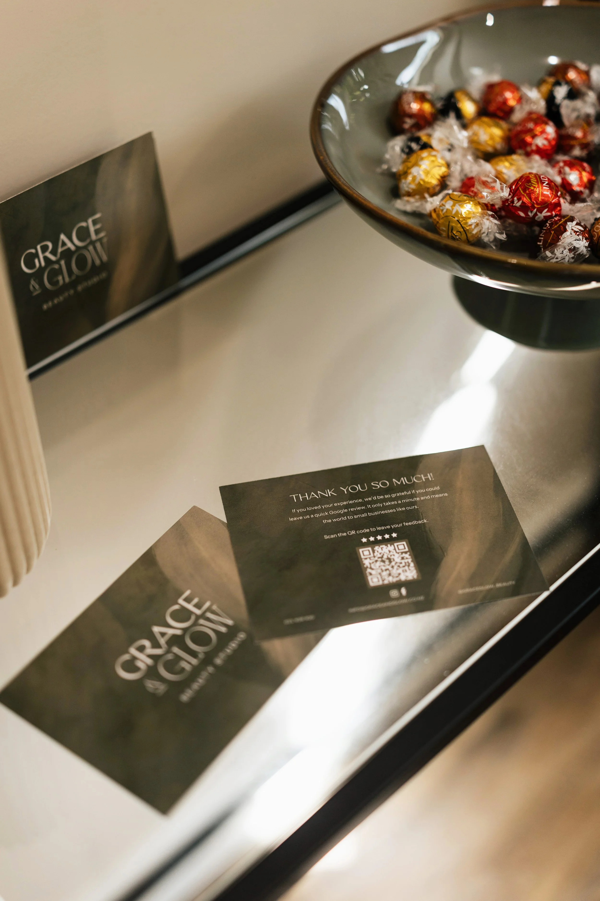 A bowl of colorful wrapped candies on a silver tray, with a 'Grace & Glow' card and a thank you card with a QR code on a reflective surface.