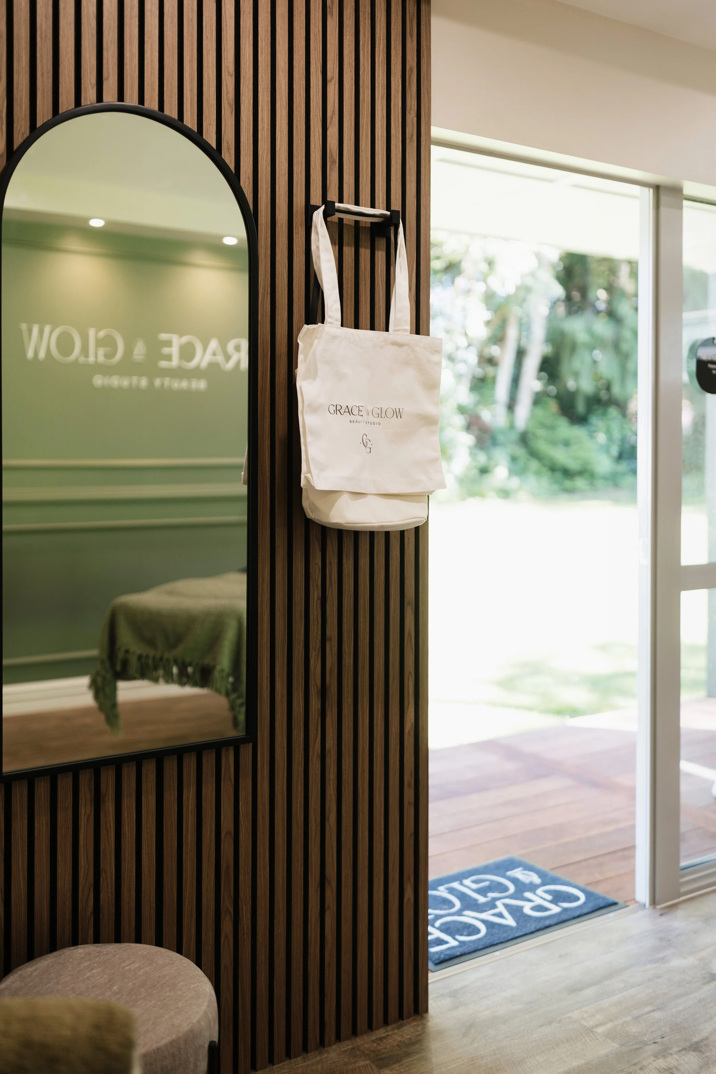 A white tote bag with the logo 'GRACE & GLOW' hangs on a black hook on a wooden slat wall near a door with a glass panel. Inside the room, there's a mirror reflecting a green blanket on a bed or bench. Outside the door, there is a blue rug with white lettering and a view of greenery.