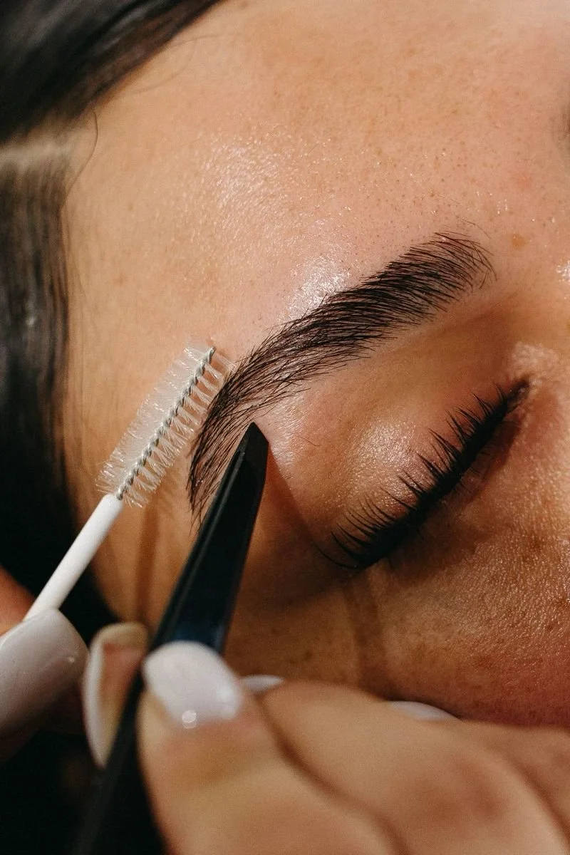 A person getting their eyebrows groomed with a spoolie brush and tweezers.
