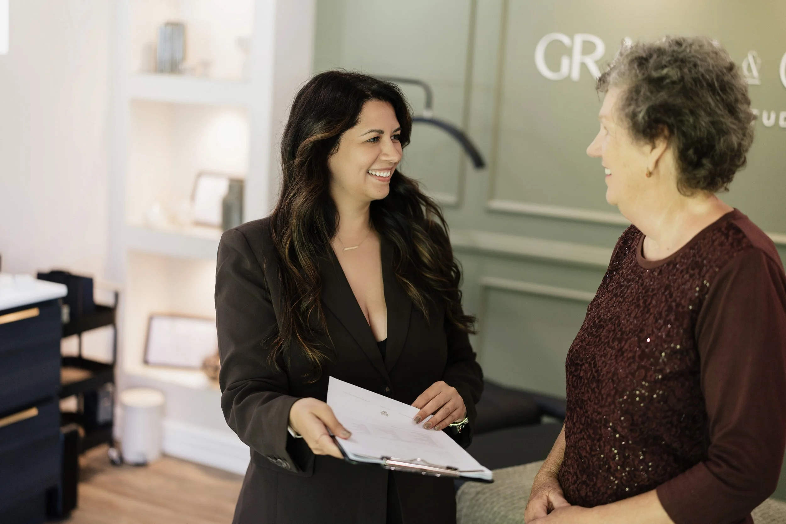 Two women having a conversation indoors, one is holding a clipboard, both are smiling. One woman has long dark hair and is wearing a black blazer, the other has short gray hair and is wearing a maroon top with sequins.