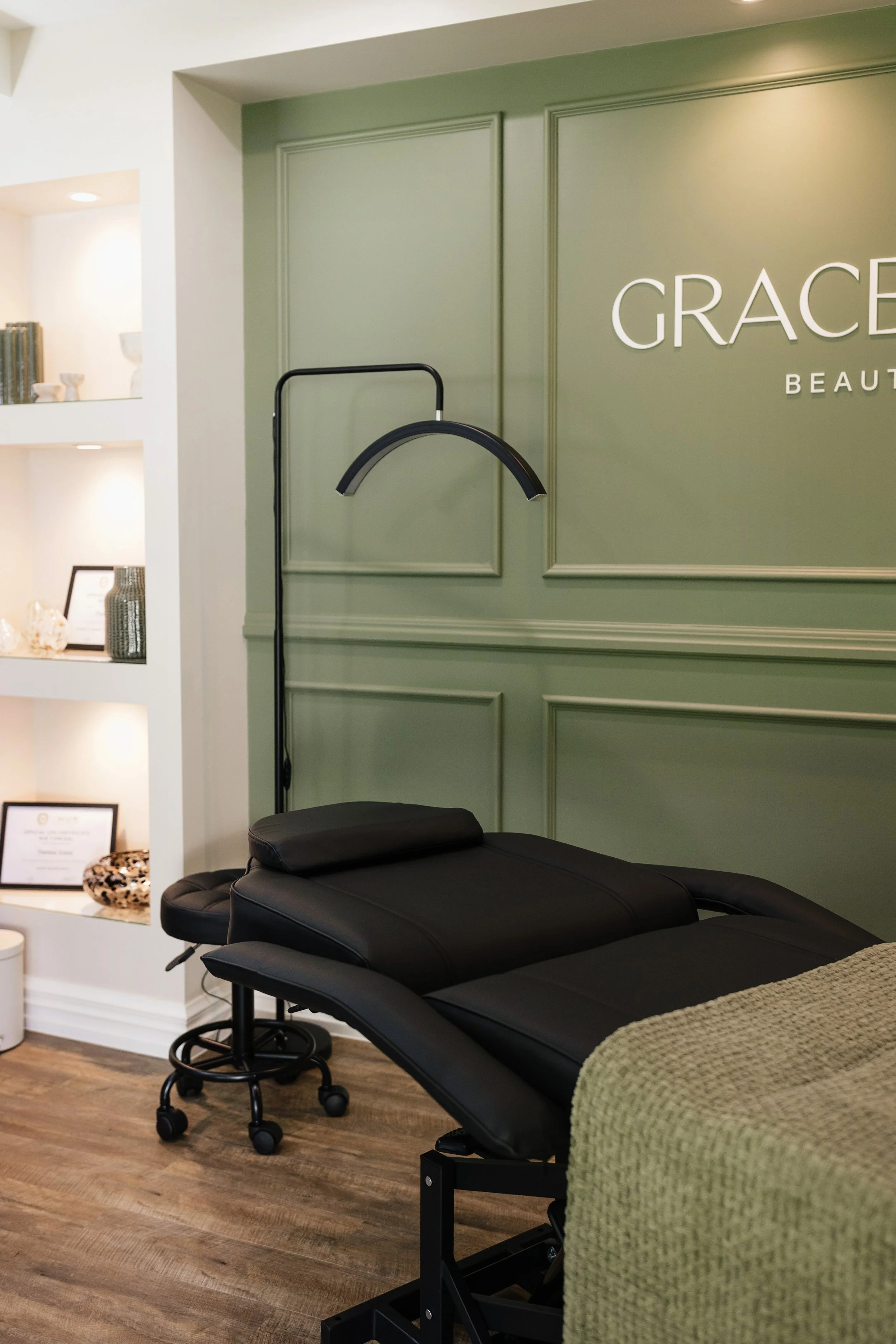 A massage or treatment room with a black treatment bed, a black rolling stool, and a green wall with the words 'GRACE BEAU' visible, part of a spa or beauty salon.