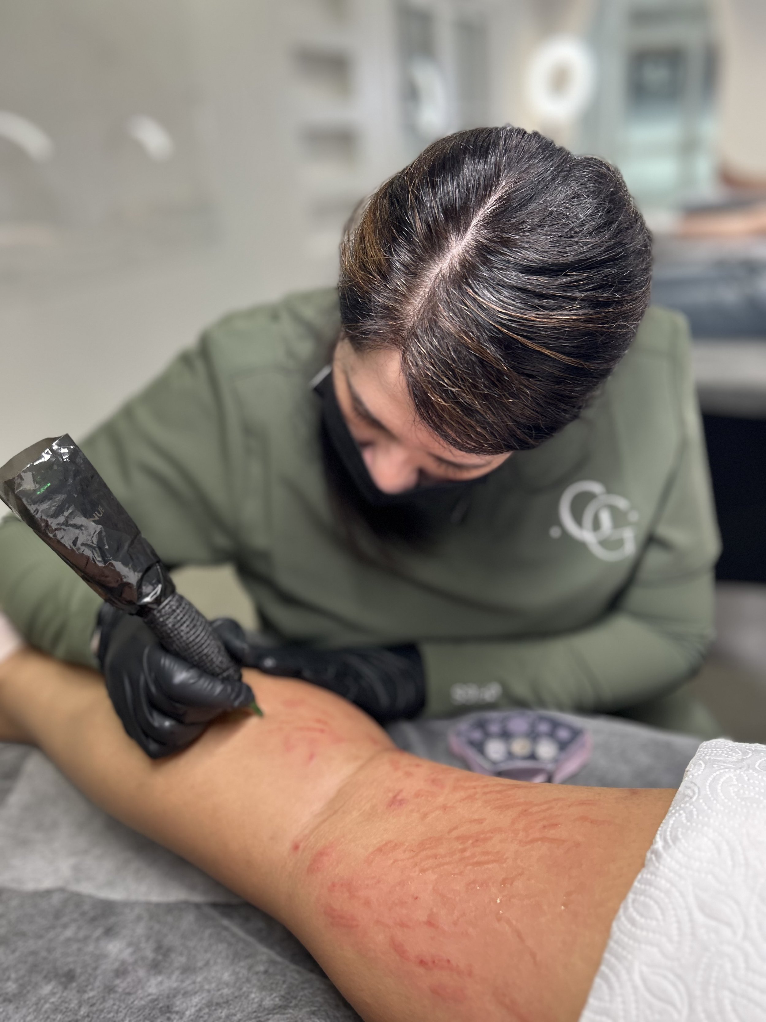 A woman in a black mask and green jacket tattoos a person's forearm with a tattoo machine. The forearm shows red, irritated skin.