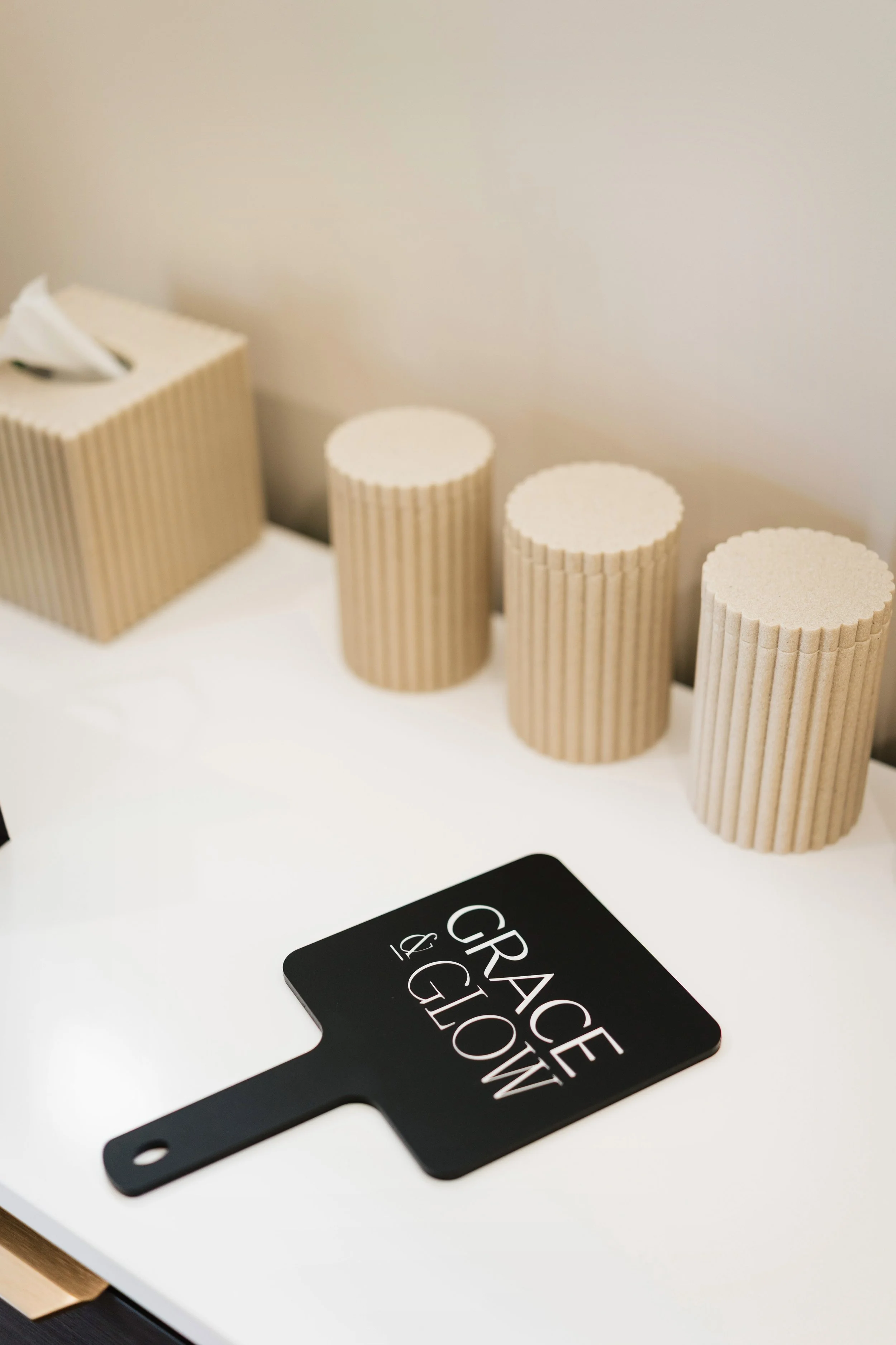 A decorative wall shelf with beige tissue box and three beige ribbed cylindrical containers, along with a black paddle-shaped sign reading 'Grace & GLOW' in white text on a white table.
