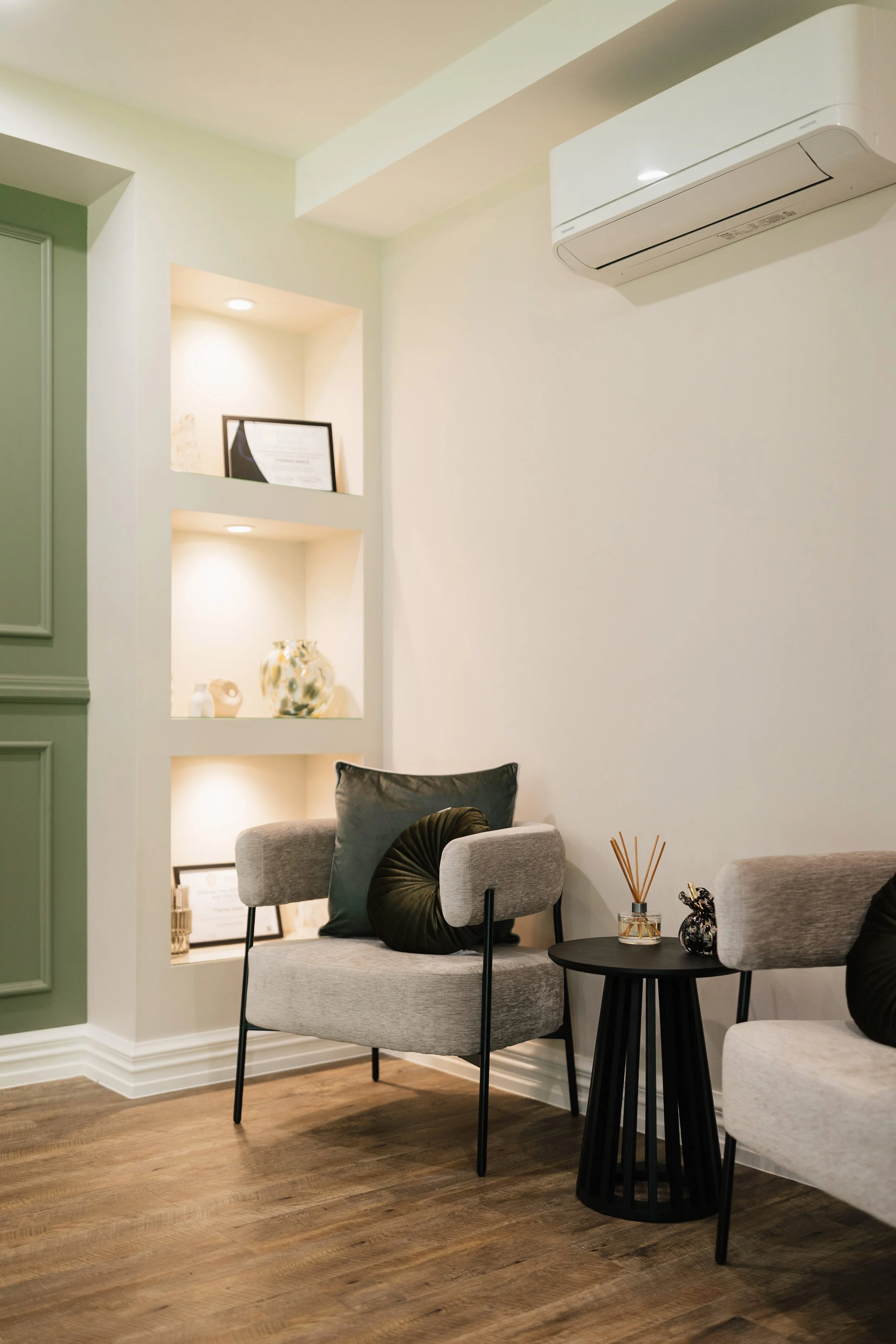 A cozy living room corner with two light gray chairs, black accent pillows, a black side table with decorative items, a built-in wall shelf with illuminated display niches, and a white wall-mounted air conditioner.
