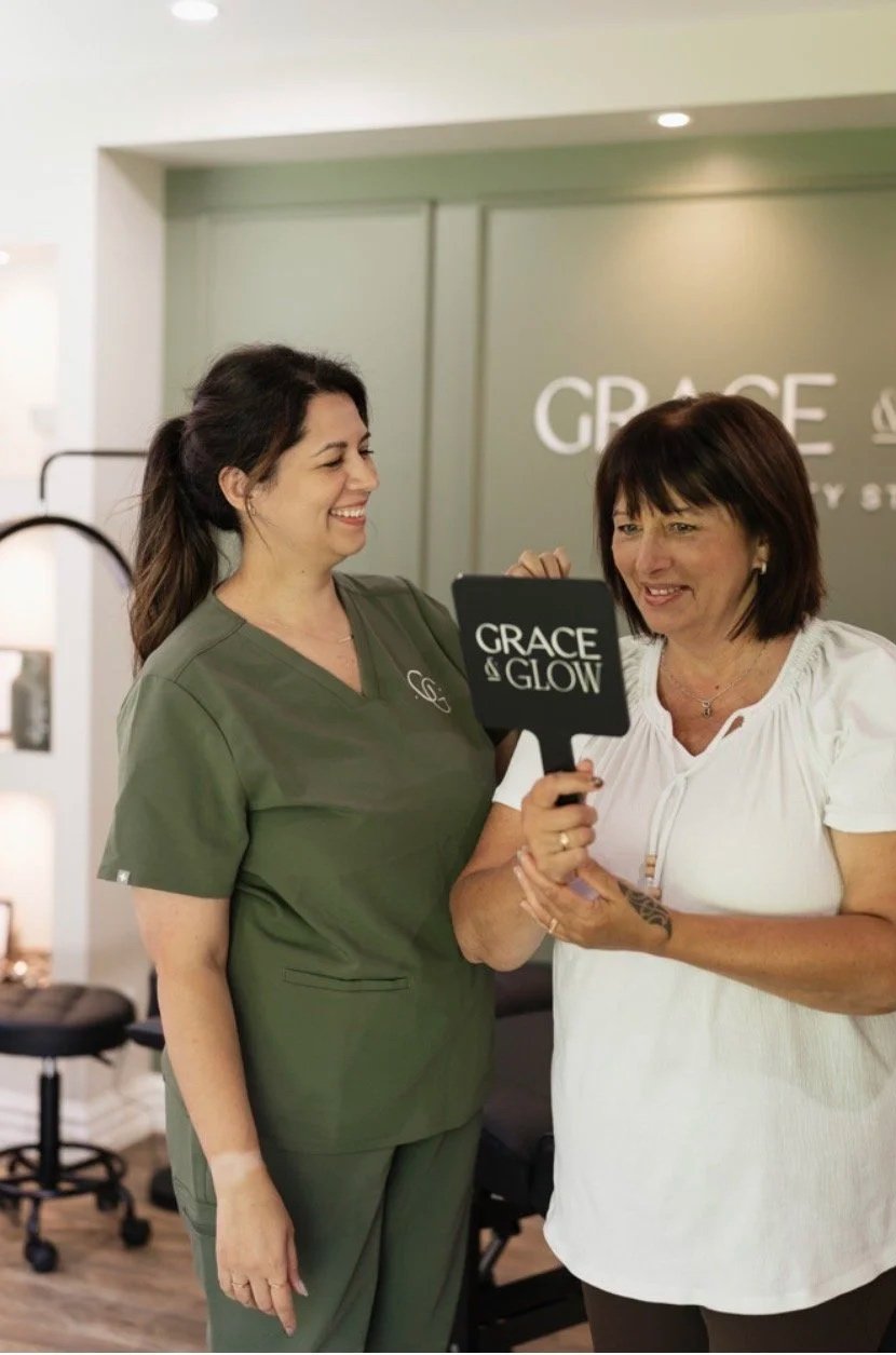 Two women smiling and holding a black fan with the words 'GRACE & GLOW' written on it inside a room with a grey wall in the background.