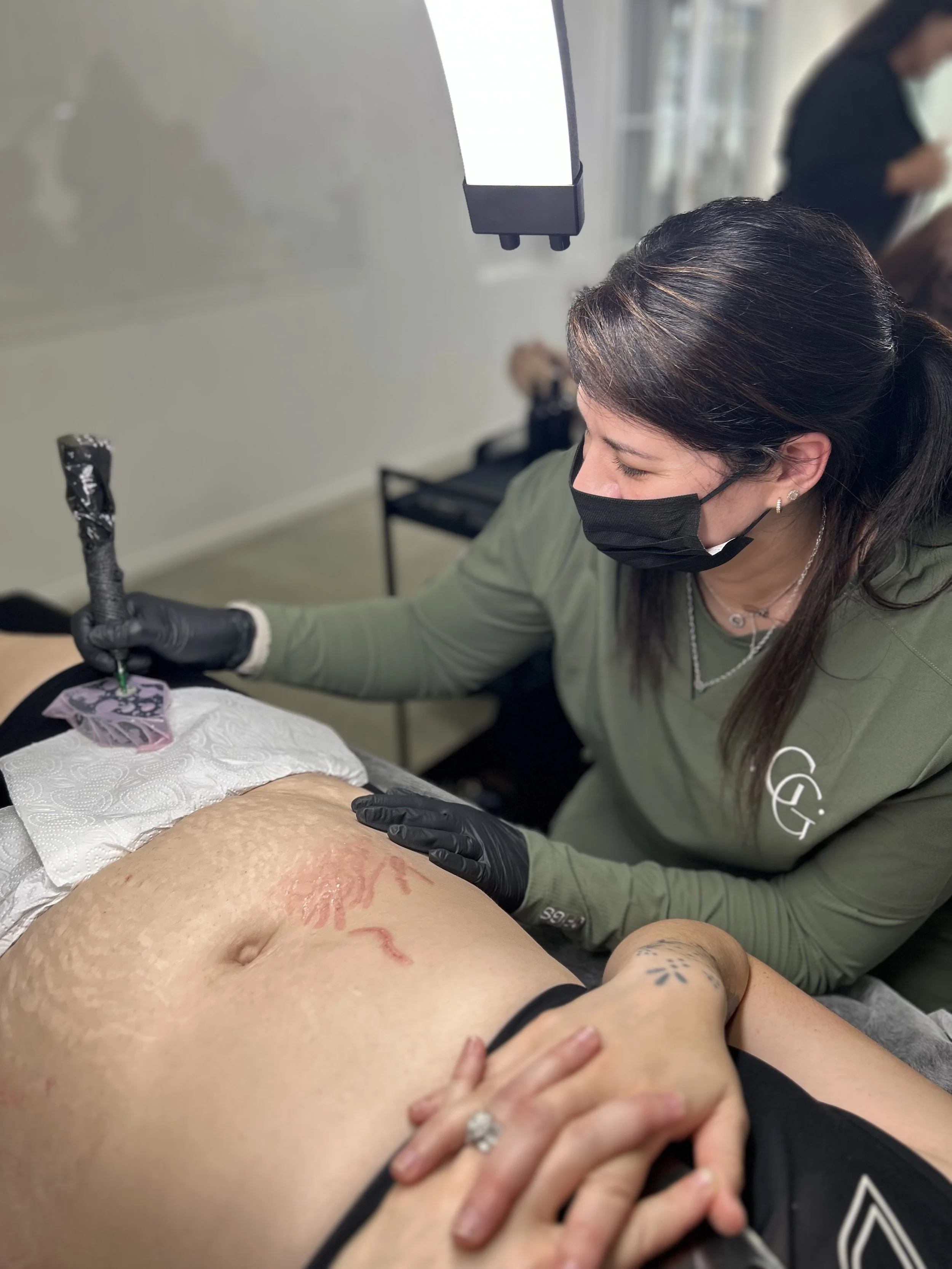 A woman with dark hair, wearing a black face mask and black gloves, is performing a tattoo on a person's abdomen. The person is lying down with their hand resting on their stomach. The tattoo artist is using a tattoo machine, and the person's skin appears to be slightly red from the tattooing process.