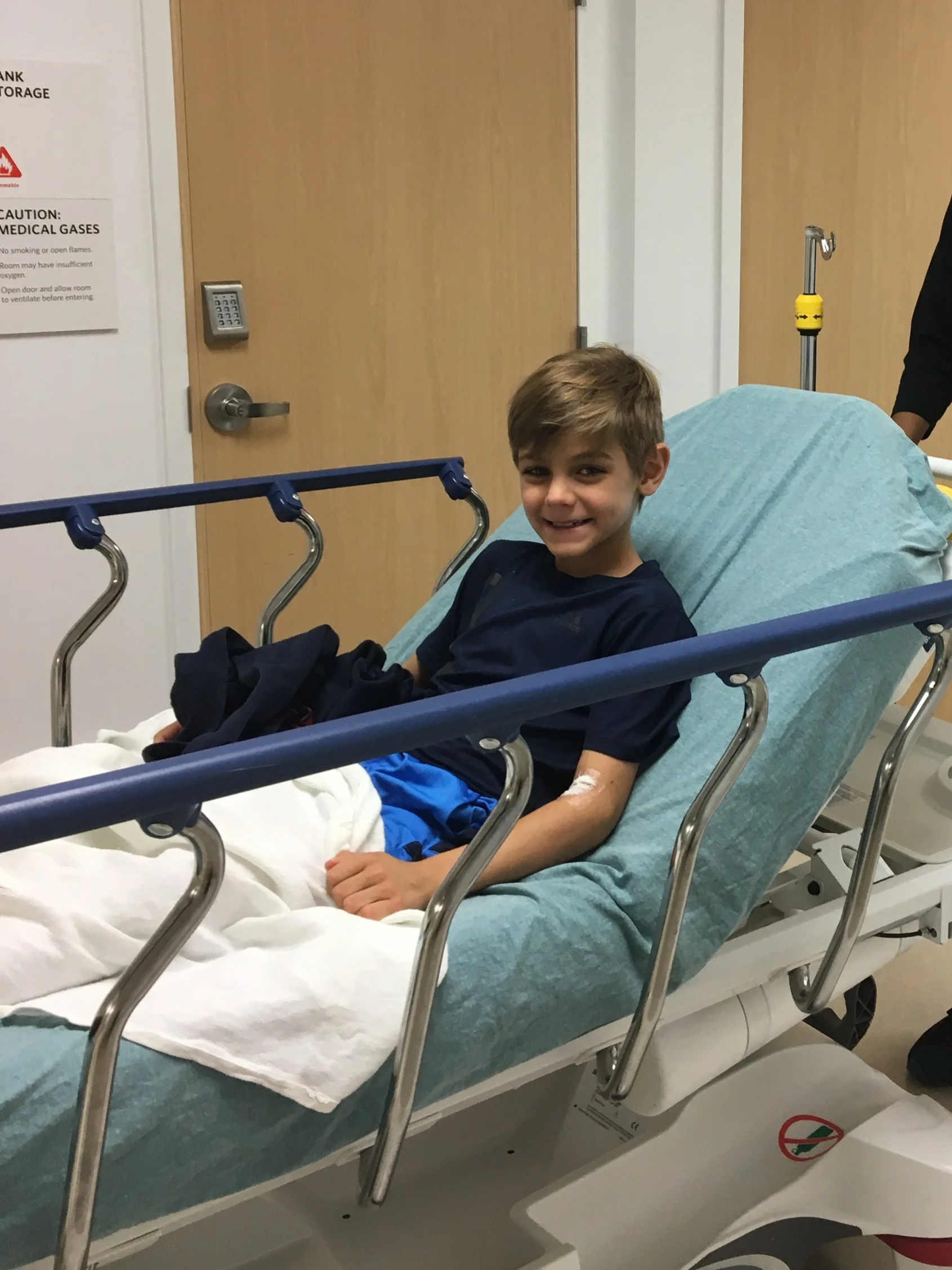 A young boy lying on a hospital bed, smiling, with medical supplies and equipment in the background.