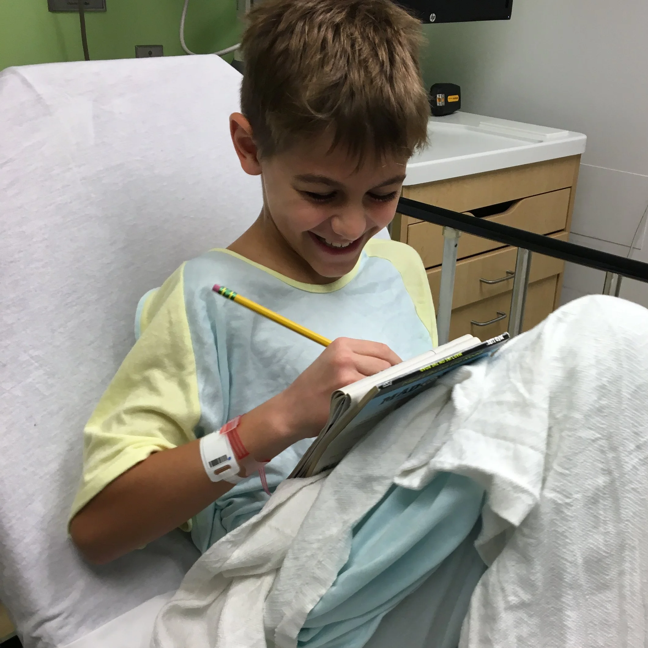 A young boy with blond hair smiling and writing with a pencil on a notepad at a hospital bed, wearing a light yellow hospital gown and a hospital wristband. The room has a white surface and a wooden drawer, with a green wall and hospital equipment in the background.