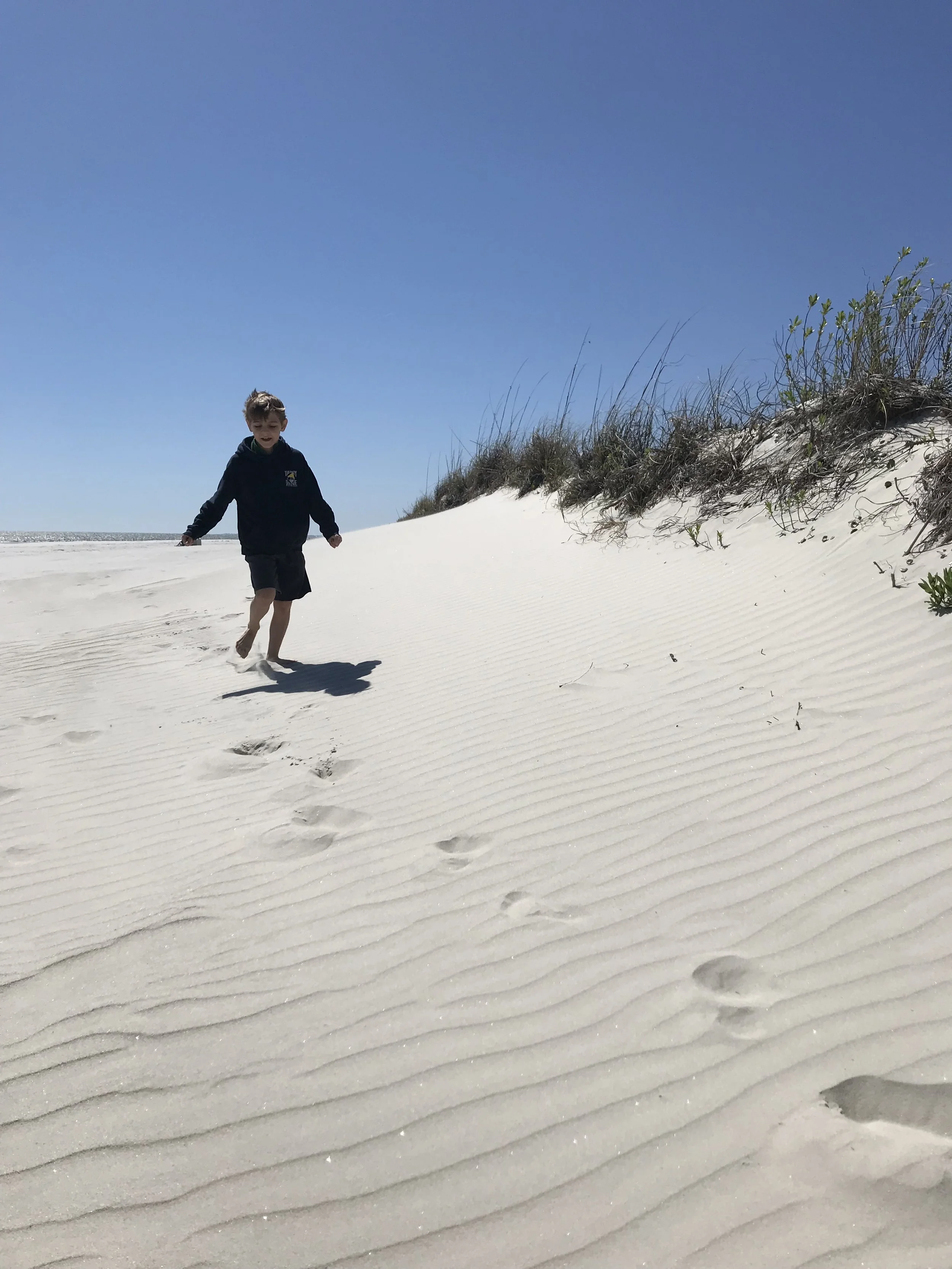 A boy running on white sandy beach with footprints, dunes with grass, and a clear blue sky.