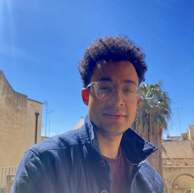 A man with curly hair and glasses stands outdoors on a sunny day with palm trees and buildings in the background.