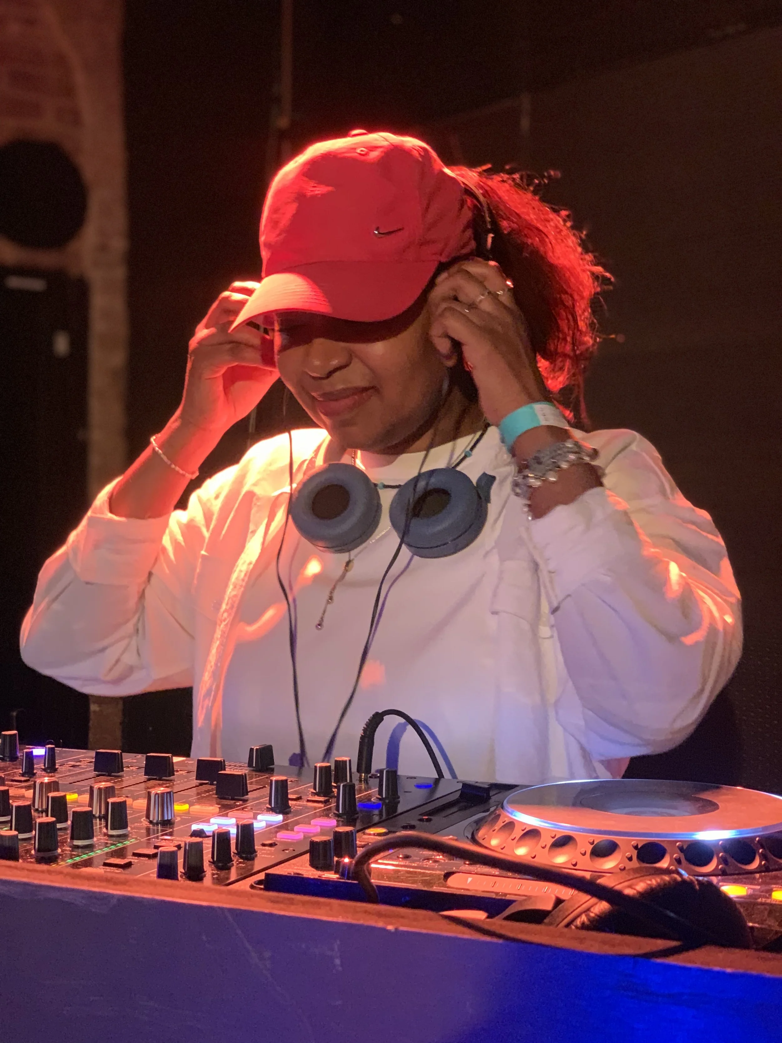 A woman DJ wearing a red cap, headphones, and a white jacket, operating a DJ mixer with colorful lights.