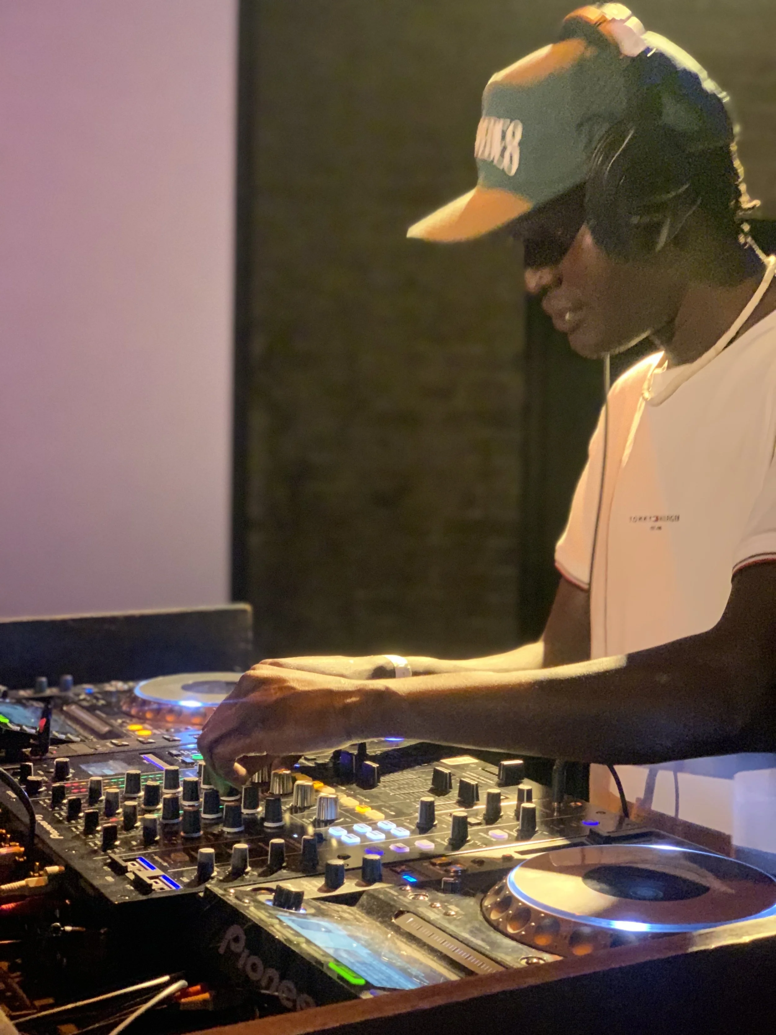 A DJ wearing a cap, sunglasses, headphones, and a white shirt with red trim, is operating a professional DJ mixer and turntables in a dimly lit setting.