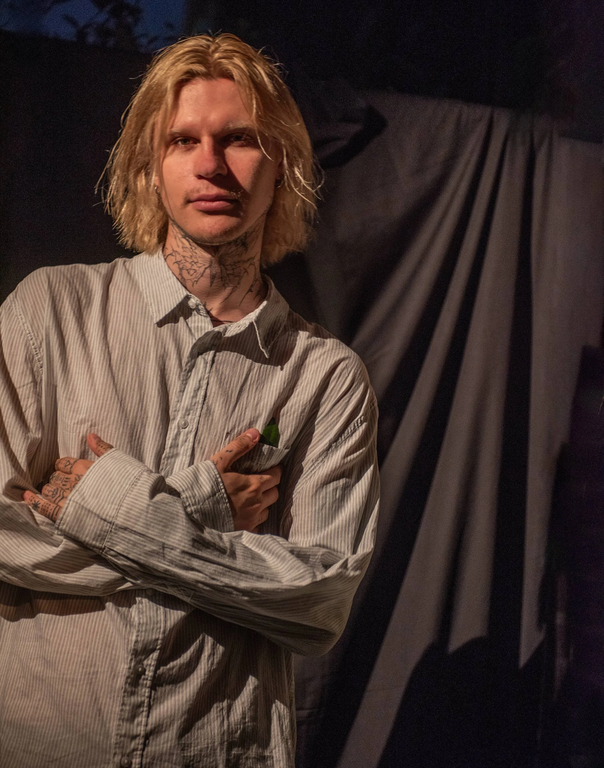 A young man with blond hair, tattoos on his neck and hands, wearing a striped button-up shirt, standing in front of a dark background with draped fabric, looking at the camera.