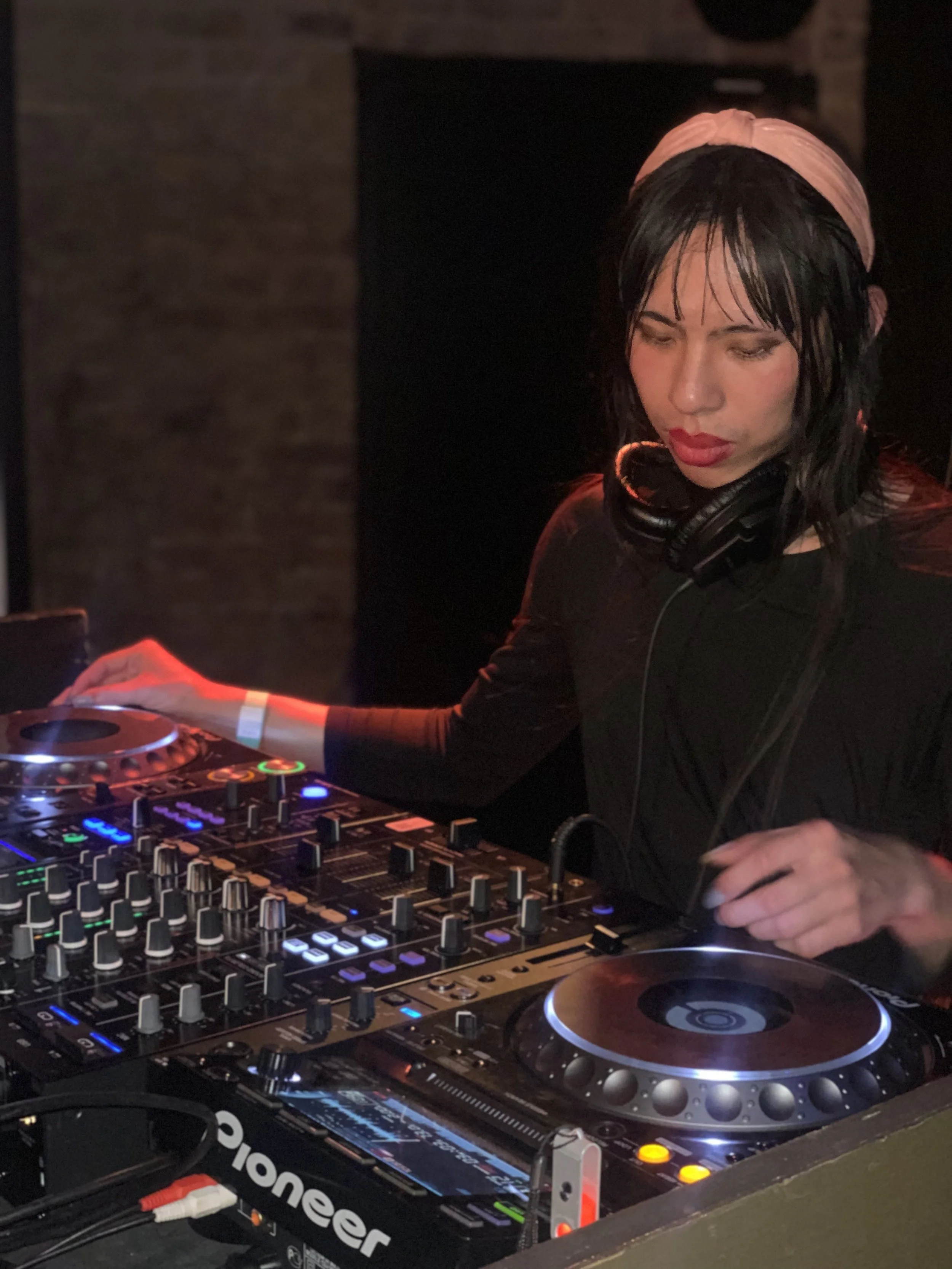 A female DJ with dark wet hair, wearing a pink headband, mixing music on a Pioneer DJ controller in a dimly lit environment.