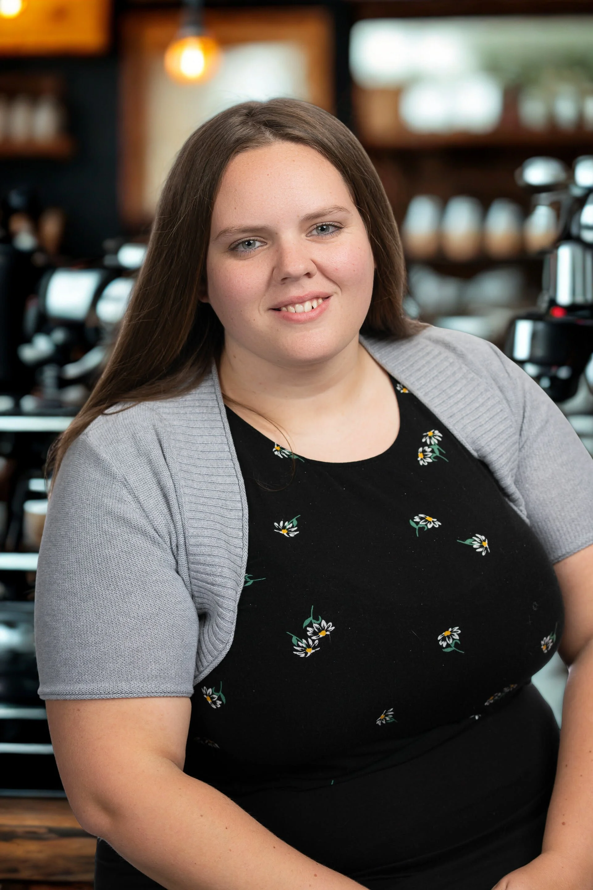 A woman with long brown hair, wearing a black dress with white daisies and a gray cardigan, smiling in a coffee shop.