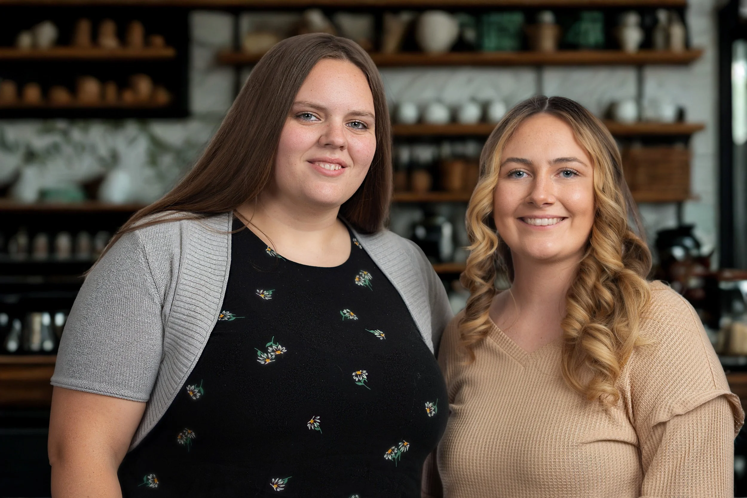 Two women standing inside a cafe or bakery, smiling at the camera, with shelves of jars and pottery in the background.