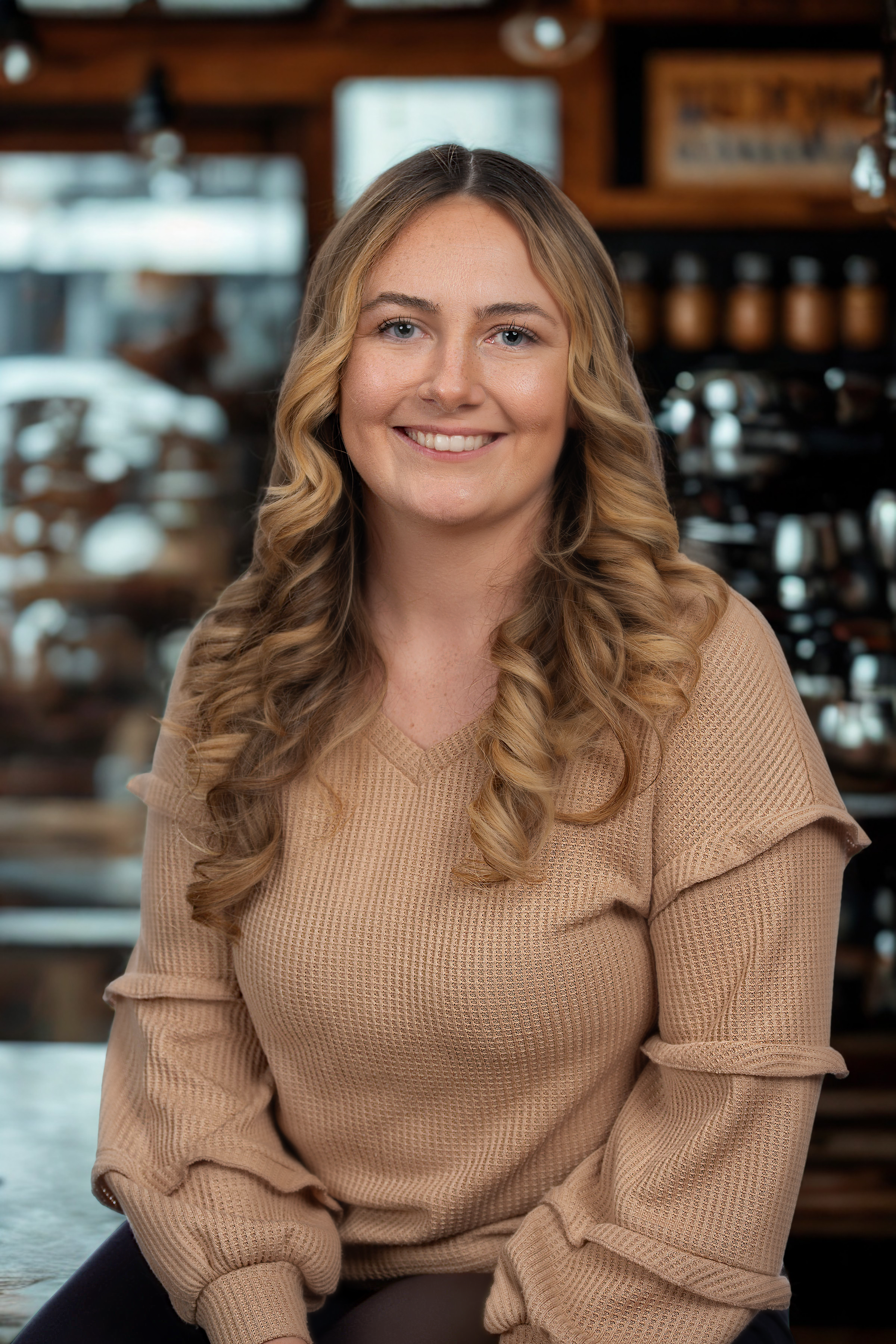 A woman with long blonde curly hair smiling, sitting in a cozy cafe or bakery with a wooden interior and shelves filled with ceramics behind her.