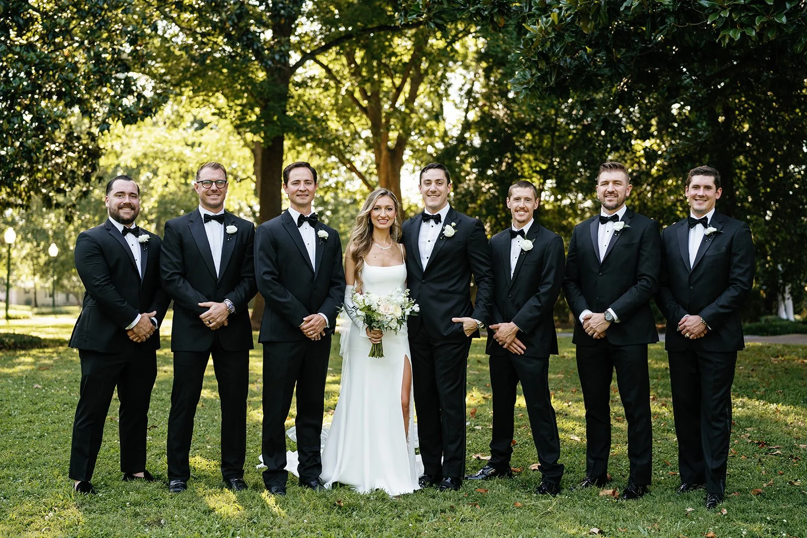 A wedding party standing outdoors on grass with trees in the background. The bride is in the center holding a bouquet, wearing a white dress, and the groom is next to her in a black tuxedo. Six groomsmen in black tuxedos are standing beside them, thr