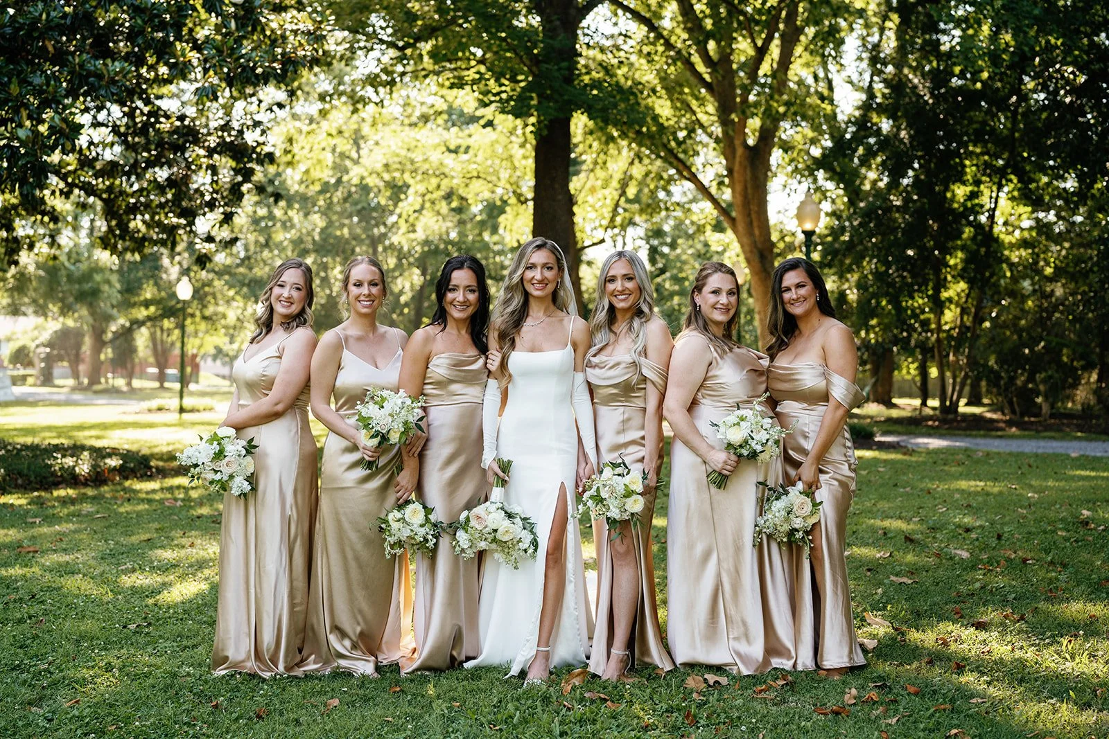A bride and six bridesmaids standing outdoors in a park, dressed in formal dresses, holding bouquets of white flowers, with green trees and sunlight in the background.