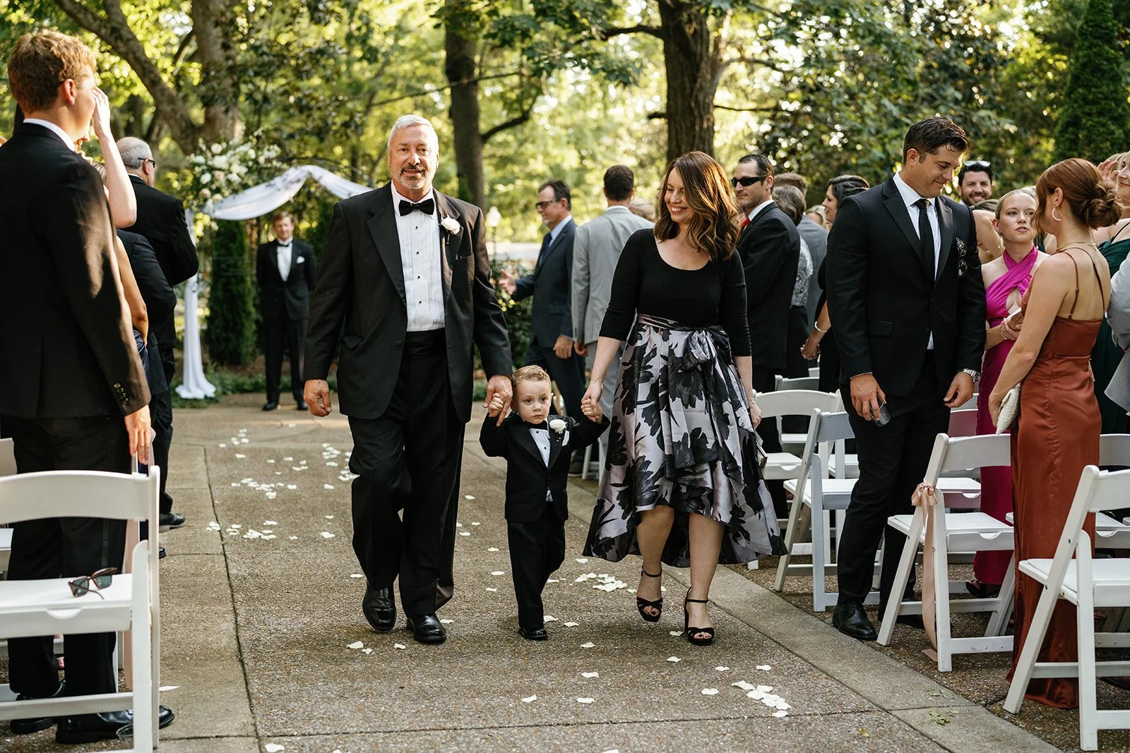 A wedding procession with a young boy, man, and woman walking down an outdoor aisle lined with chairs, surrounded by guests, trees, and wedding decorations.