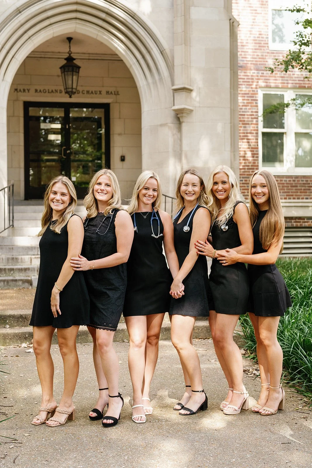 Six Vanderbilt graduates in black dresses with stethoscopes around their necks standing outside a building with steps and arched entrance, smiling at the camera.