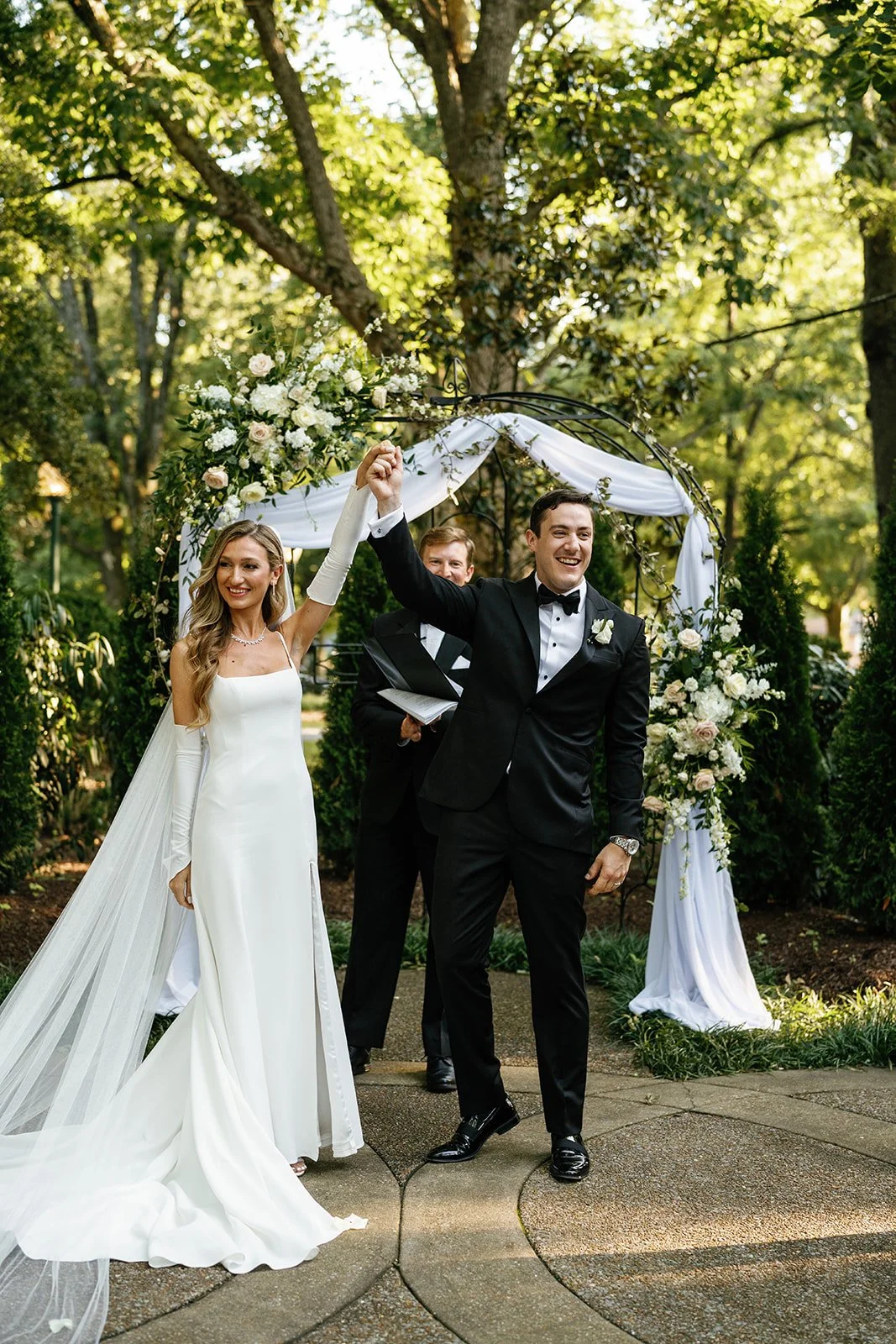 A newlywed couple celebrating their wedding ceremony outdoors under a floral arch, with the groom in a black tuxedo and the bride in a white wedding gown, holding hands and smiling.