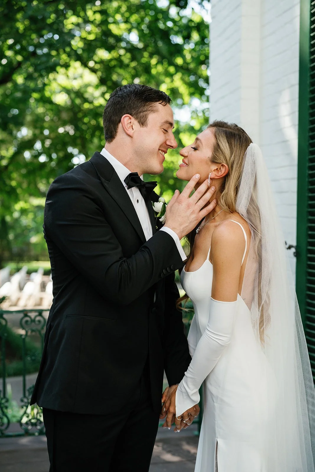 A bride and groom on their wedding day, standing close and smiling at each other, outdoors with green trees in the background. The groom is in a black tuxedo and the bride is in a white wedding dress with long gloves and a veil.