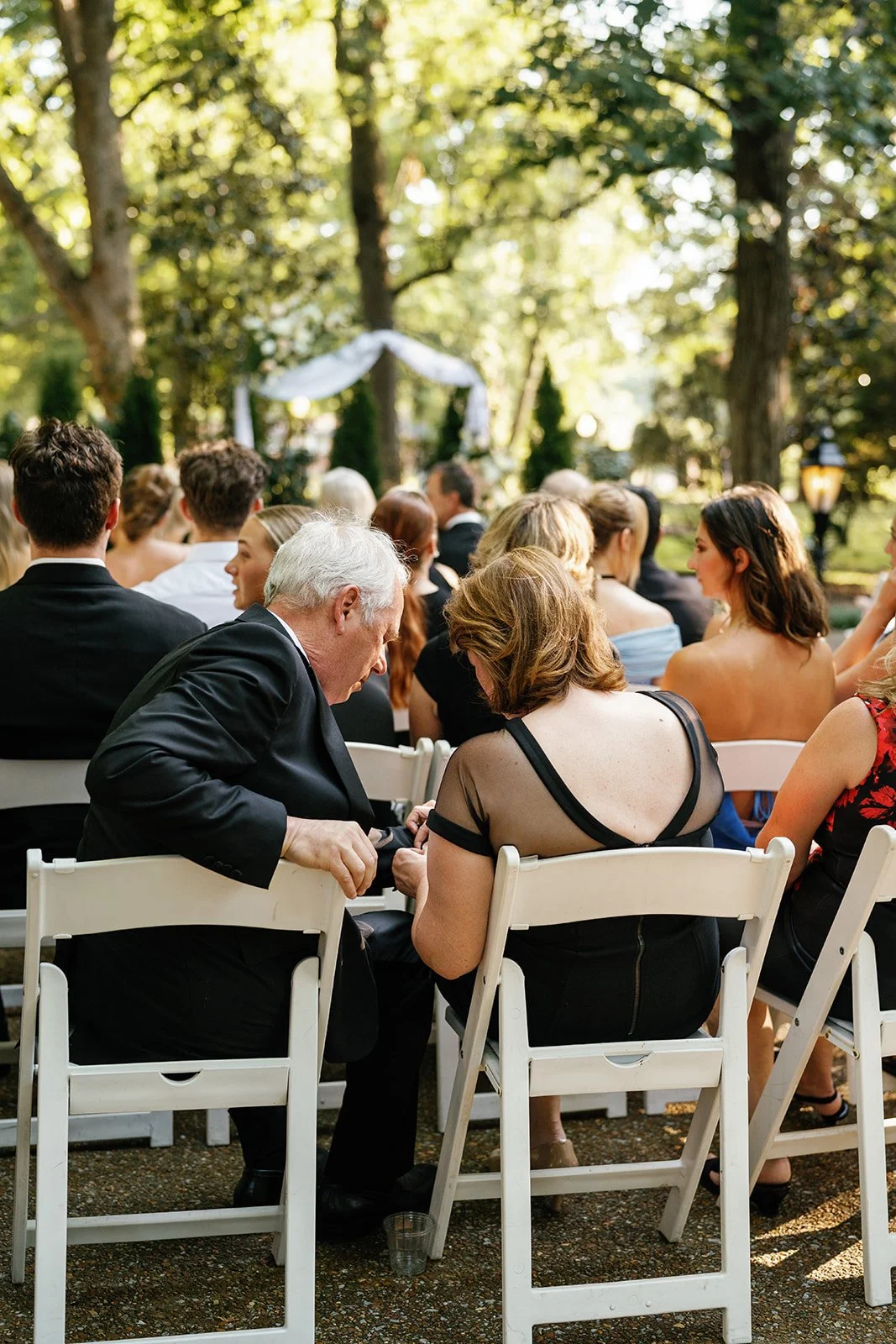 People gathered at an outdoor wedding ceremony seated in white chairs, with trees and greenery in the background.