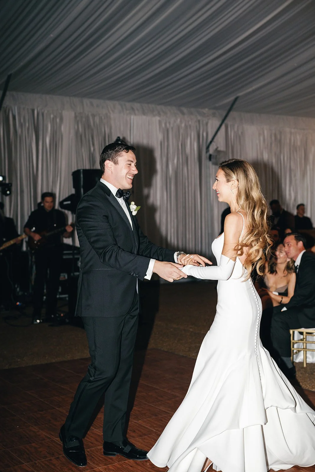 Bride and groom dancing at their wedding reception, holding hands, with guests watching in the background.