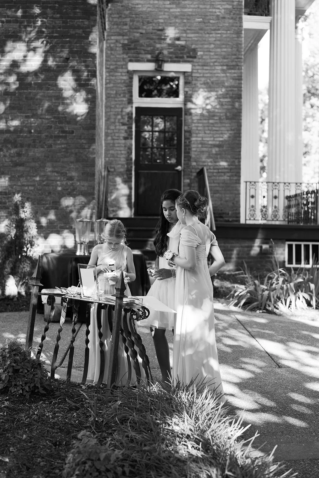 Three women and one young girl gathered around a table outside a brick building, looking at items on the table, with the scene partly shaded by trees.