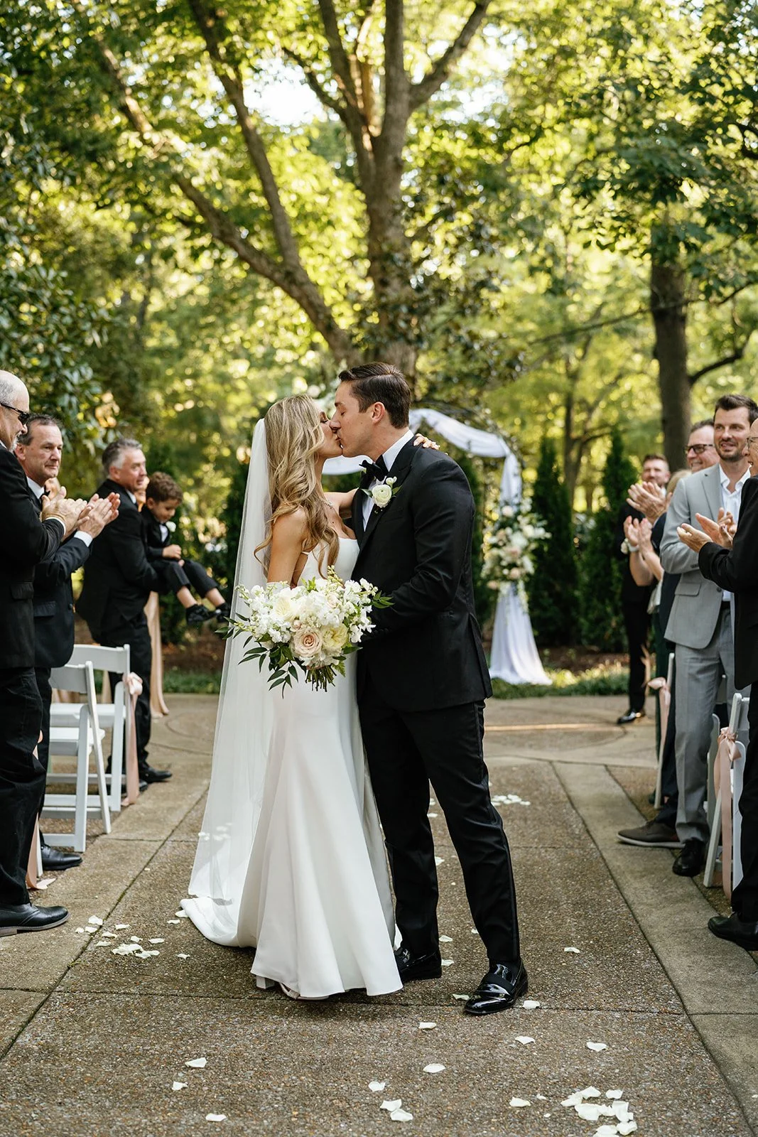 A bride and groom kiss during their outdoor wedding ceremony, surrounded by guests clapping and smiling, with lush green trees in the background.