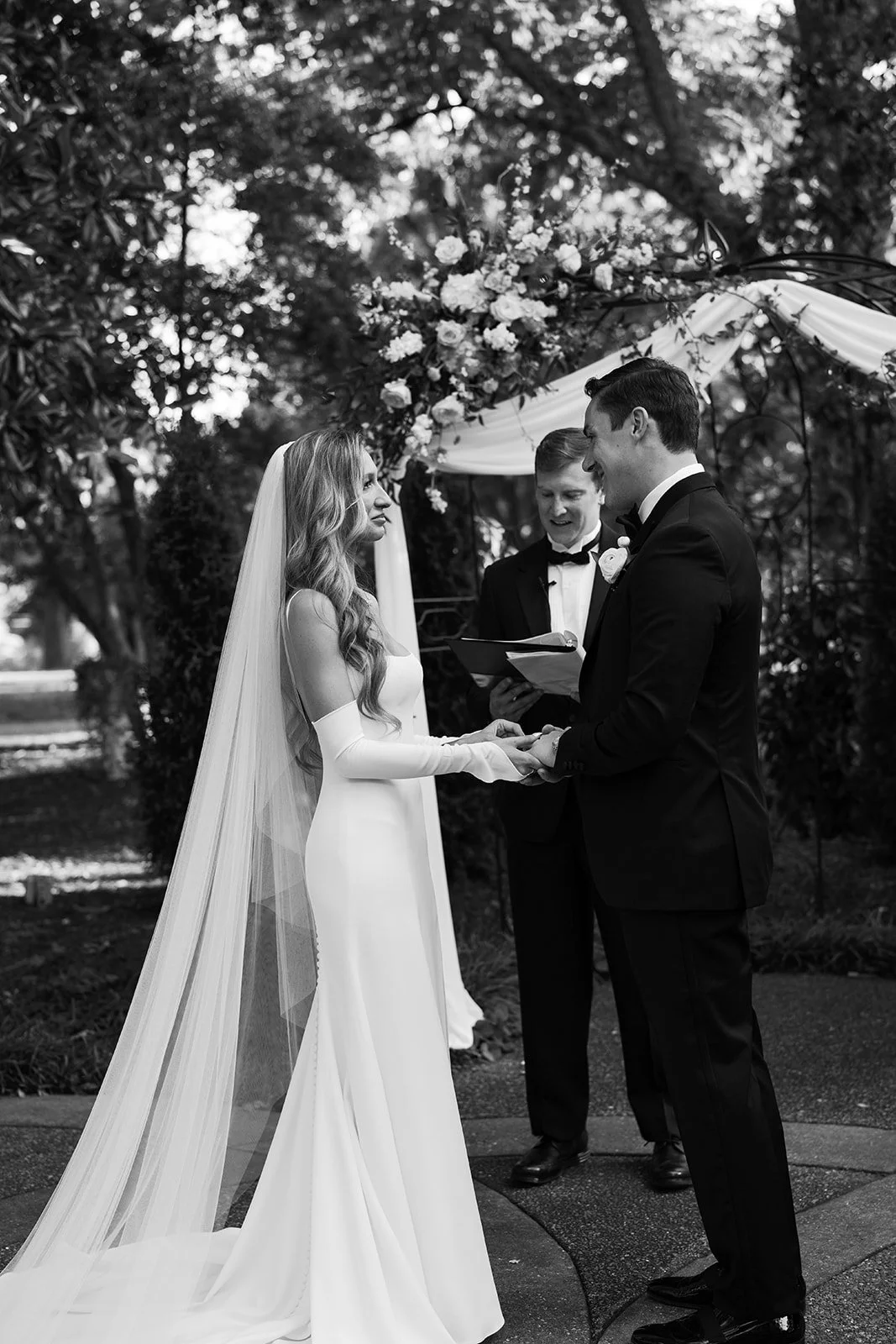 A bride and groom exchange vows during their outdoor wedding ceremony, with an officiant reading from a book, under a decorated arch of flowers and drapery, surrounded by trees.
