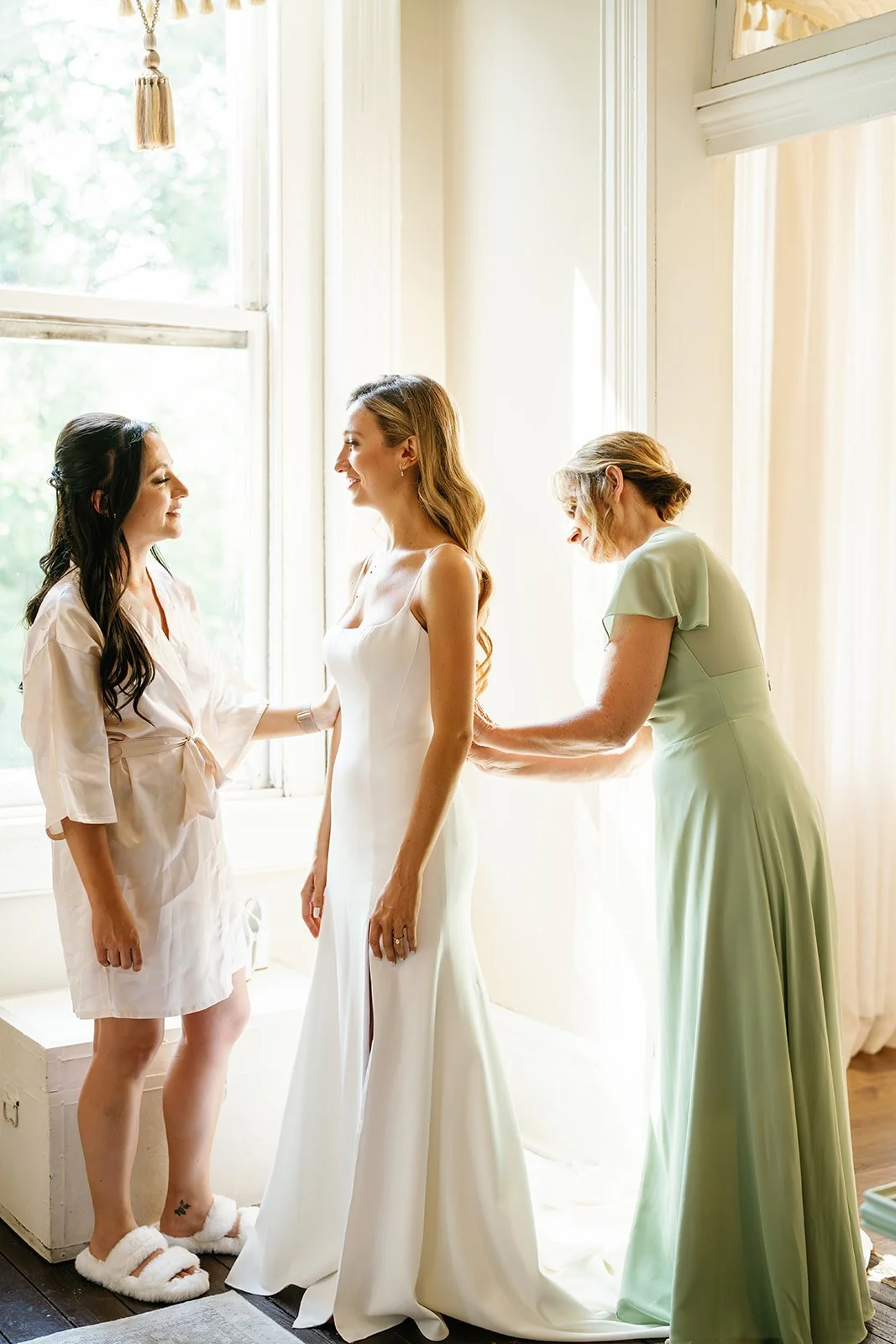 Three women near a window, with one wearing a wedding dress, another in a light pink dress, and a third in a mint green dress, preparing for a wedding.