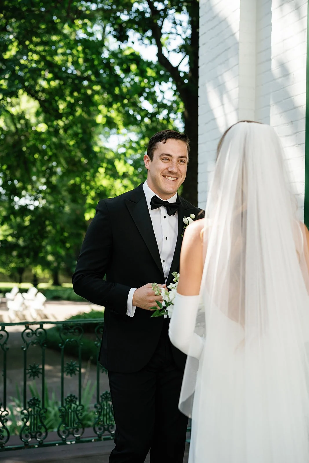 A groom in a black tuxedo with a bow tie standing outdoors, smiling at a bride who is wearing a white wedding dress with a veil, holding a bouquet of flowers.