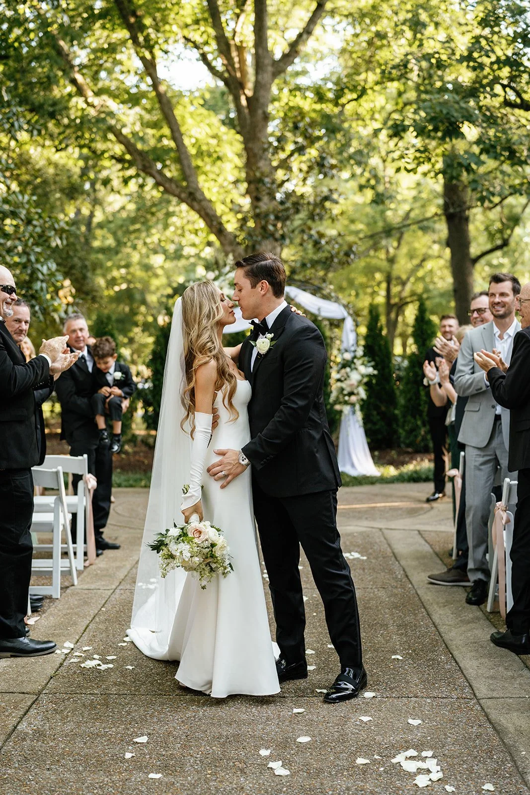 A bride and groom kiss in the middle of an outdoor wedding ceremony surrounded by smiling guests clapping, with a backdrop of trees and a decorated archway.
