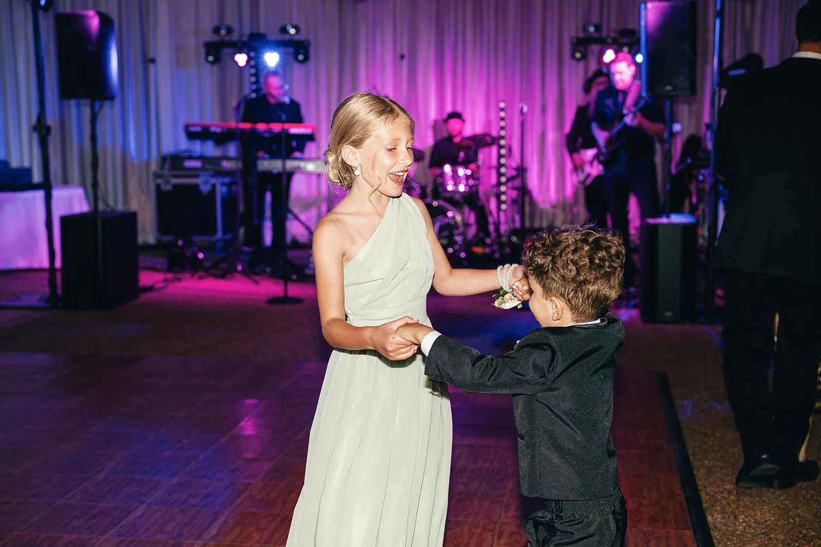 A young girl and boy dancing at a formal event with a band playing on stage in the background, decorated with purple lighting.