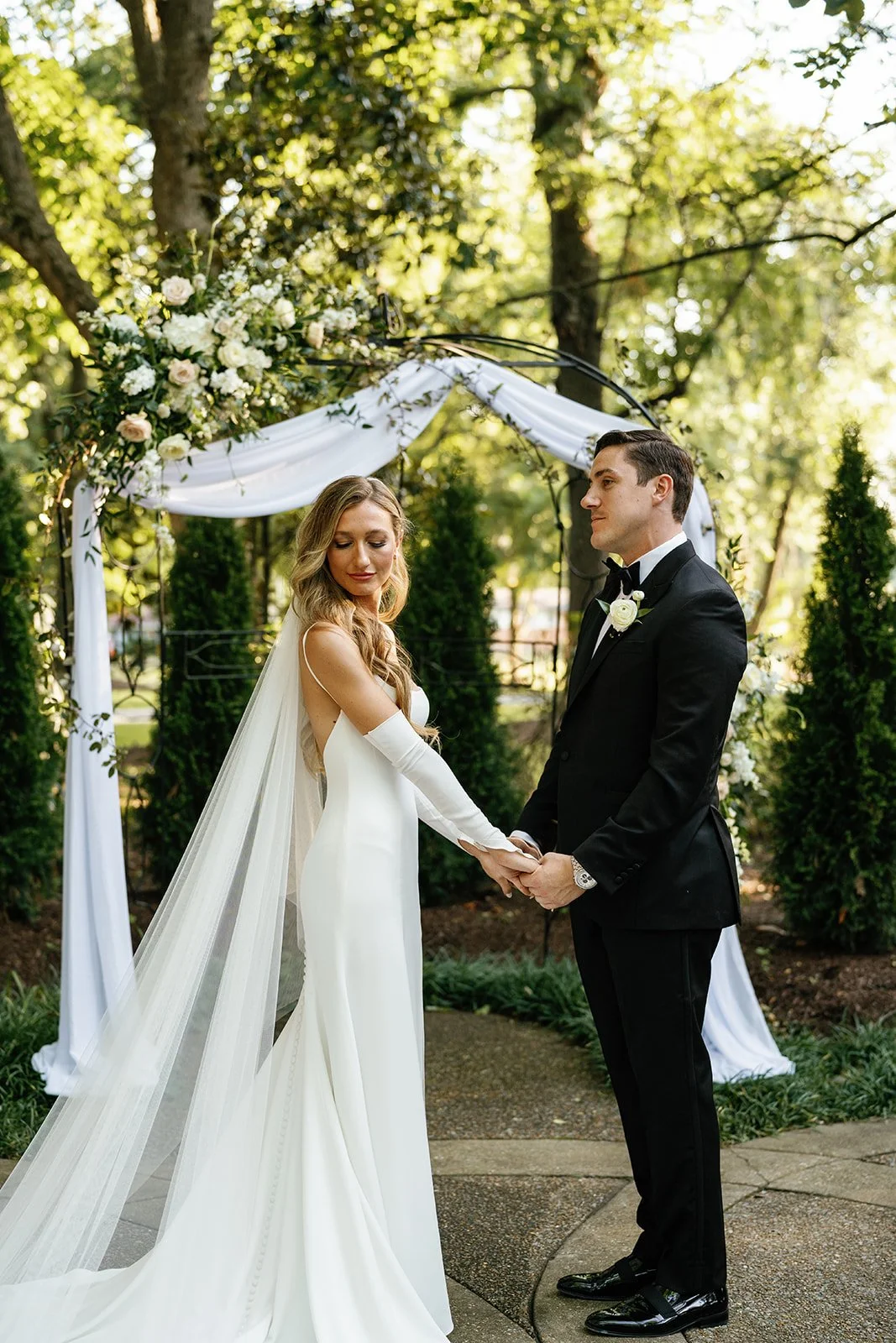 A bride and groom holding hands and looking at each other during their outdoor wedding ceremony in a garden. The bride is wearing a white wedding dress with a long veil, and the groom is dressed in a black tuxedo with a bow tie. There is a floral arc