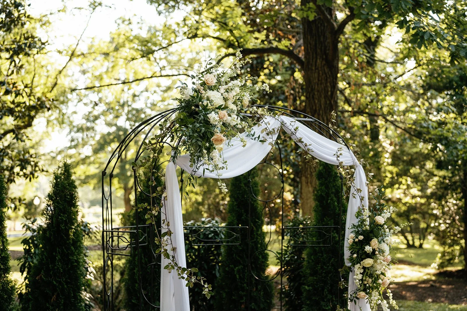 Wedding arch decorated with white flowers, greenery, and white fabric, set outdoors among trees.