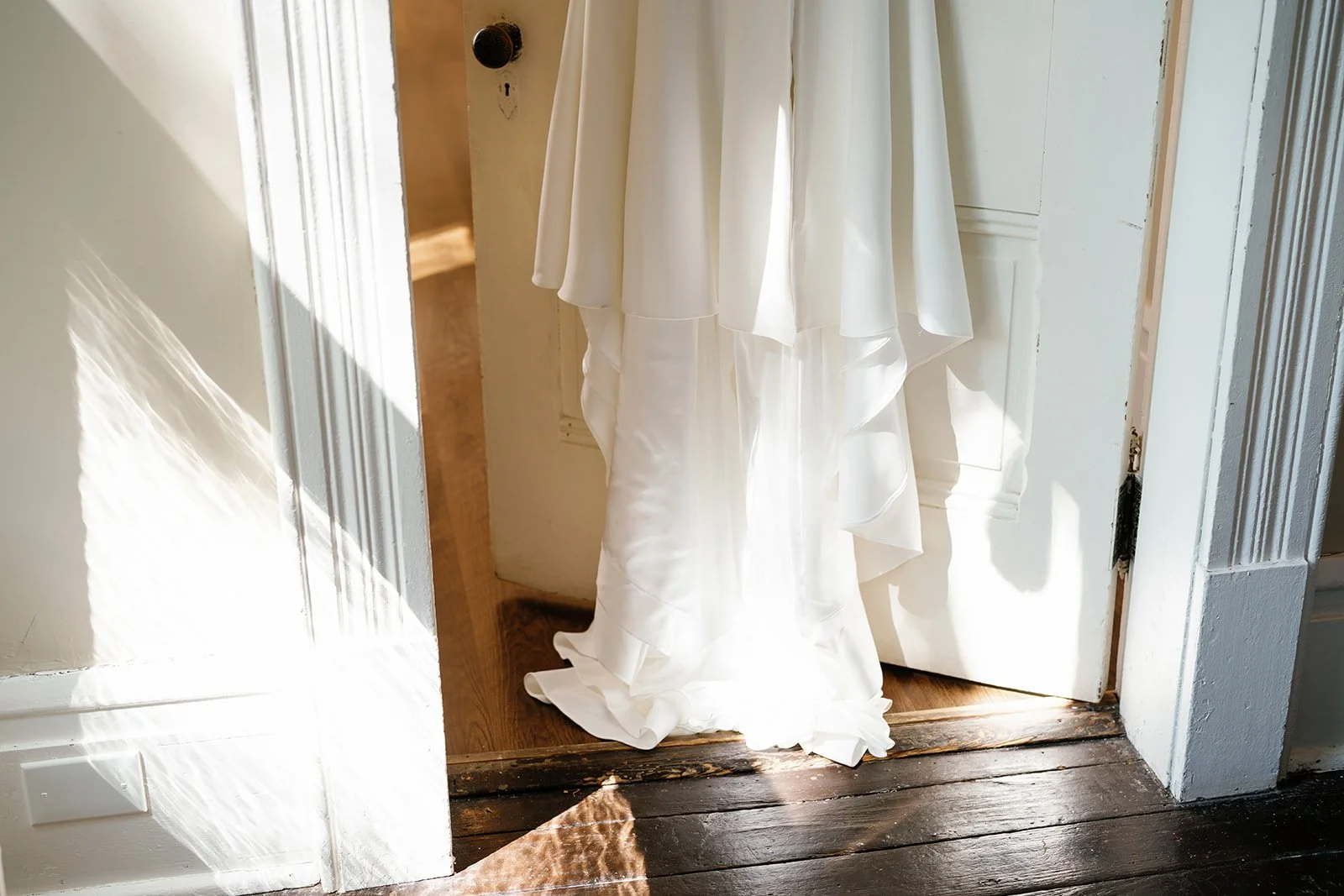 White wedding dress hanging in a doorway, illuminated by sunlight.