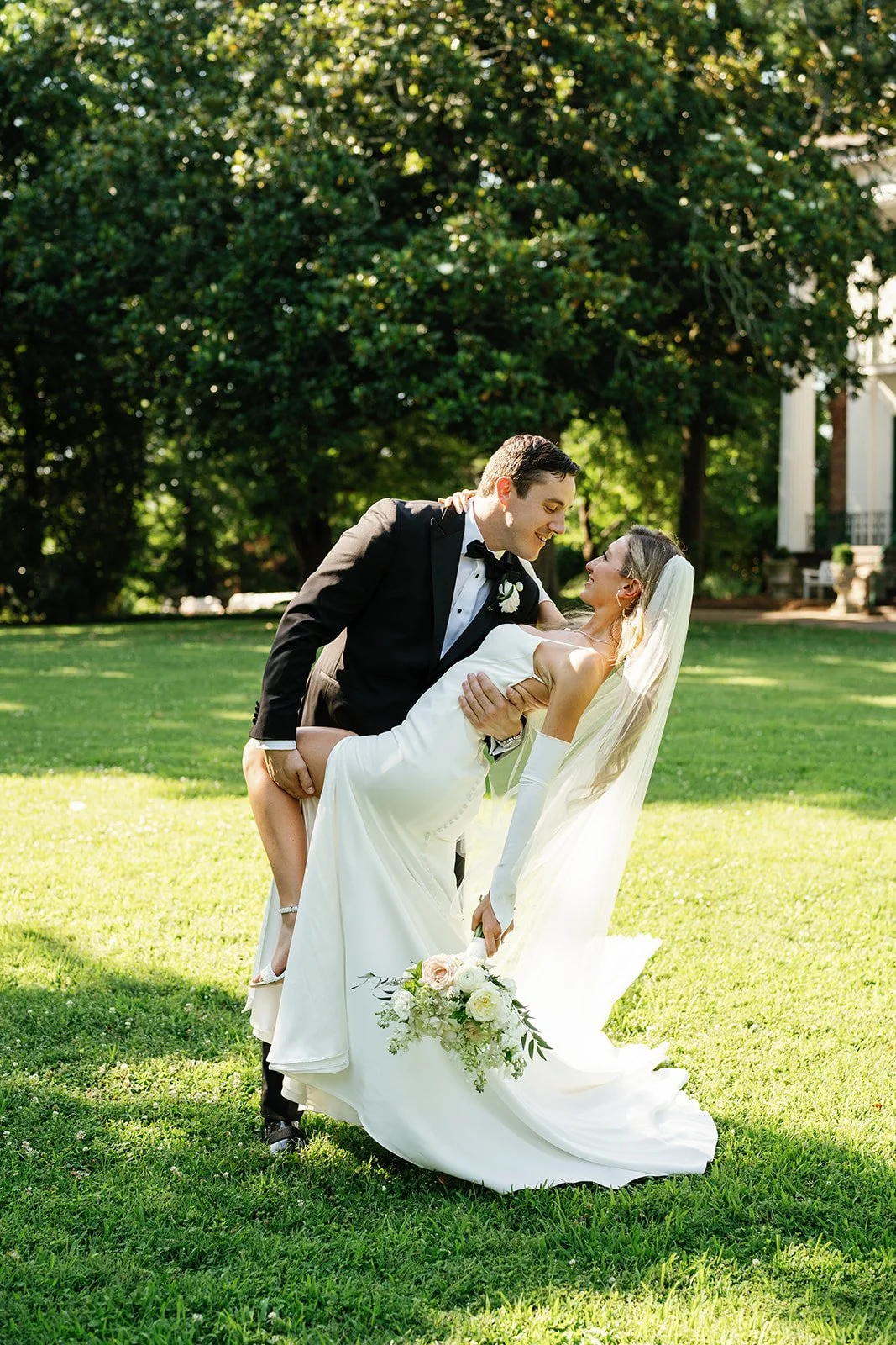 A groom in a black tuxedo holding a bride in a white wedding gown and veil in a garden with green trees and grass, smiling at each other.