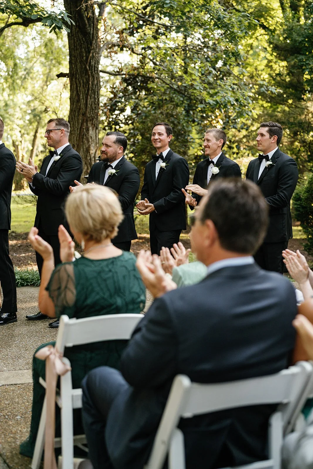 Groom and groomsmen in black tuxedos at an outdoor wedding ceremony, standing under trees, clapping and smiling, with seated guests in the foreground.