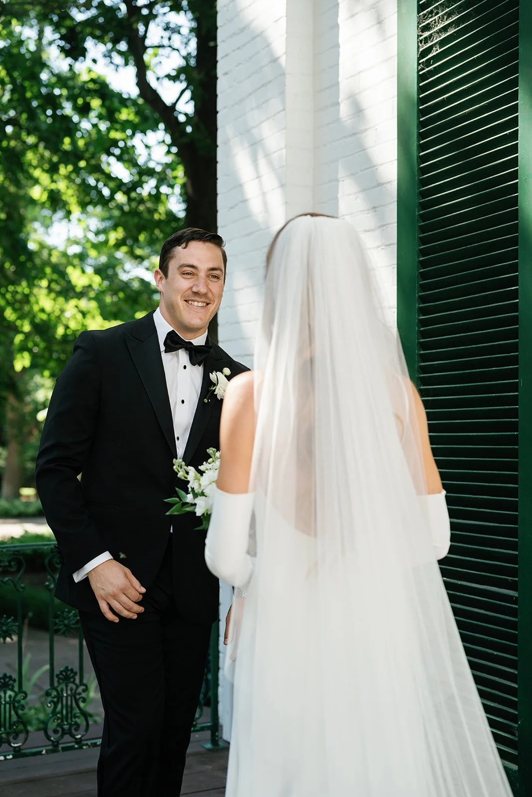 A groom in a black tuxedo and bow tie smiling at a bride in a white wedding dress and veil holding a small bouquet of white flowers outside near a white brick building with green shutters.