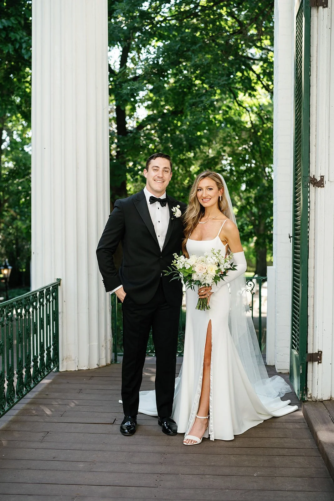 A bride and groom posing outdoors on a porch, surrounded by greenery. The groom is dressed in a black tuxedo with a bow tie, and the bride wears a white gown with a high slit, holding a bouquet of white and pink flowers.