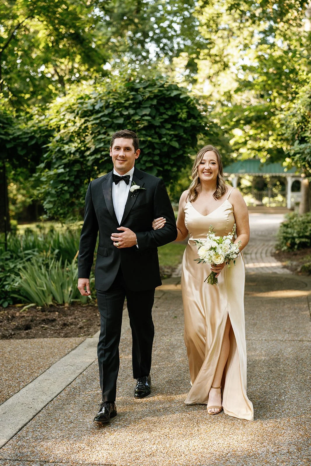 Wedding couple walking outdoors on a garden pathway, the groom in a black tuxedo and the bride in a champagne-colored gown holding a bouquet of white flowers, surrounded by lush green trees.