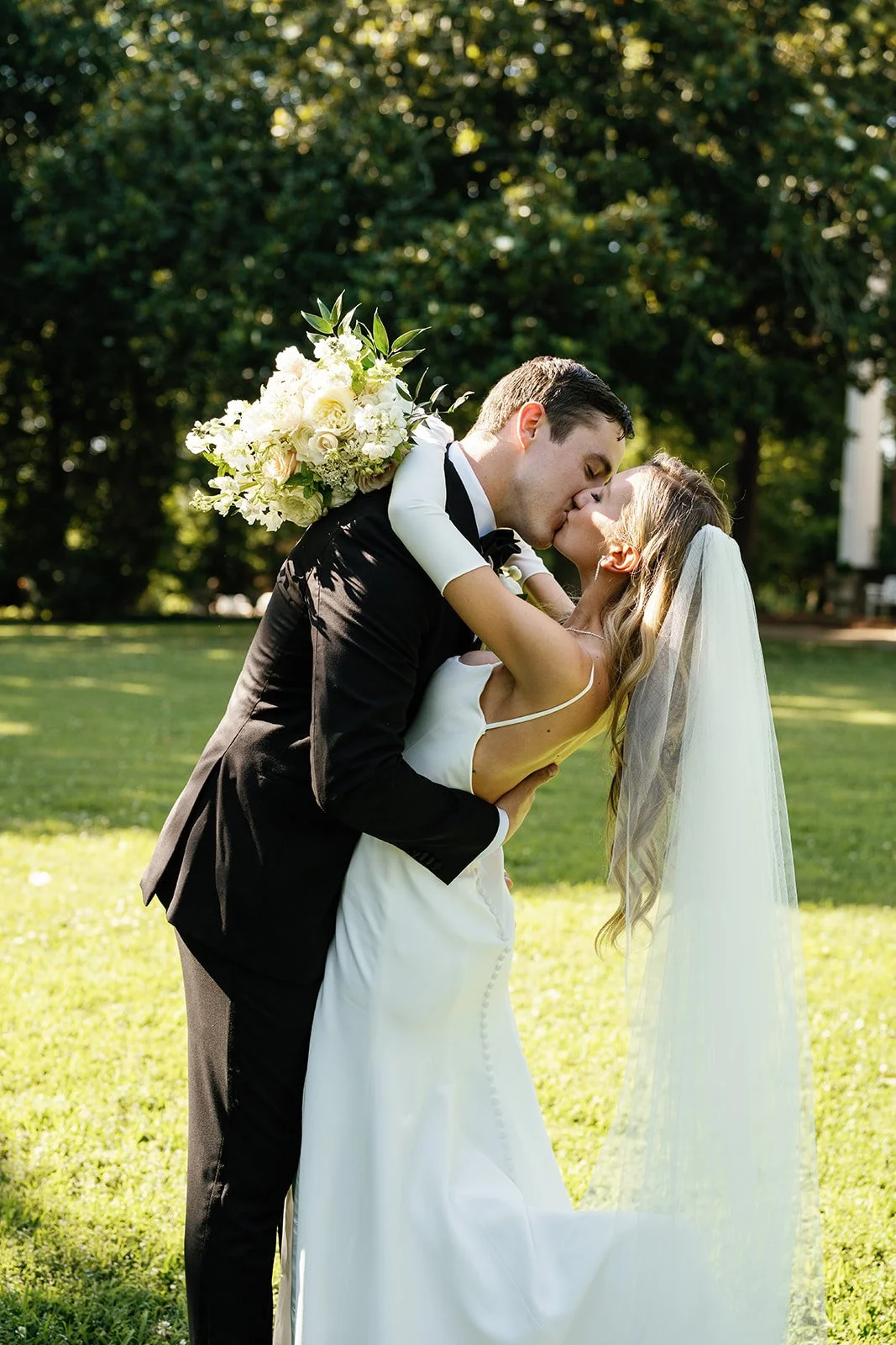 A newlywed couple sharing a kiss outdoors during daytime, the groom in a black tuxedo holding a large bouquet of white flowers, and the bride in a white wedding dress and veil, with green trees in the background.