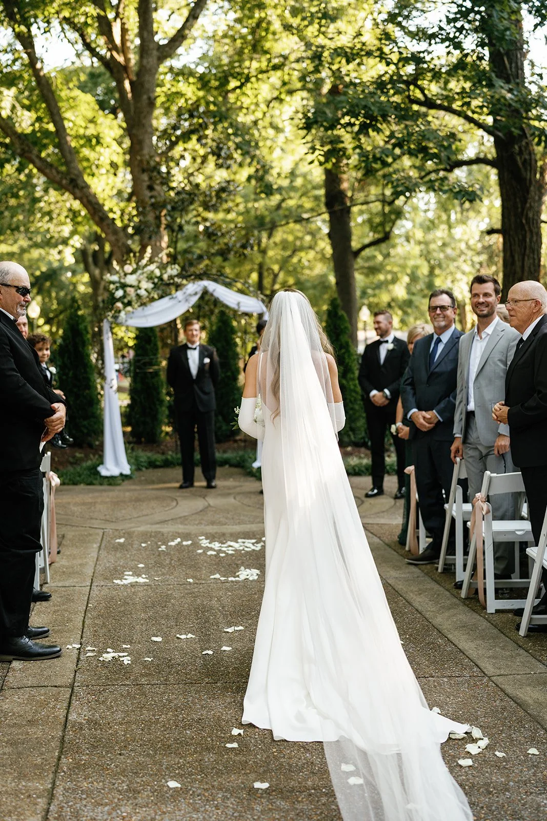 Bride in a white dress and veil walking down the outdoor wedding aisle, lined with guests on a lush green, tree-filled background.